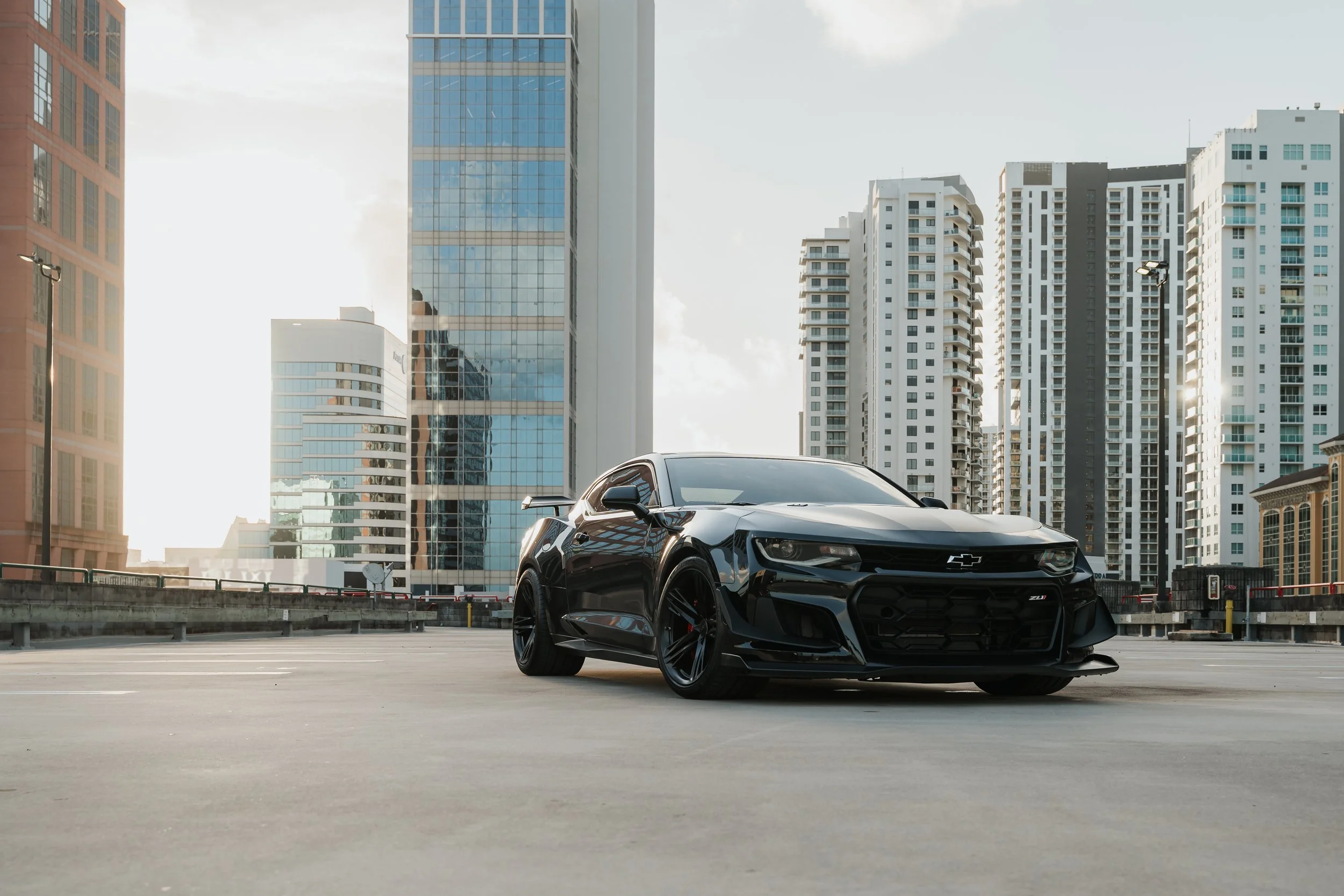 A black Chevrolet Camaro sports car parked on top of a parking garage with tall city buildings in the background.