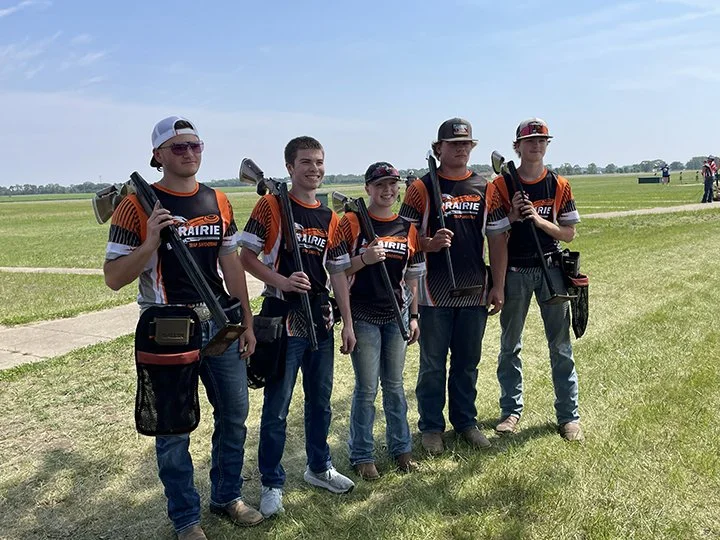 Five young men stand outdoors on a grassy field, each holding a shotgun on their shoulder, wearing matching black and orange sports jerseys, with a clear blue sky in the background