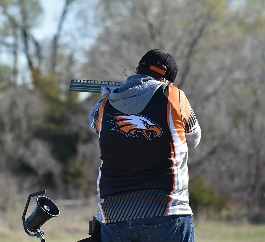 Person shooting a rifle outdoors, wearing a sports jersey with an eagle logo on the back, a baseball cap, and blue jeans.