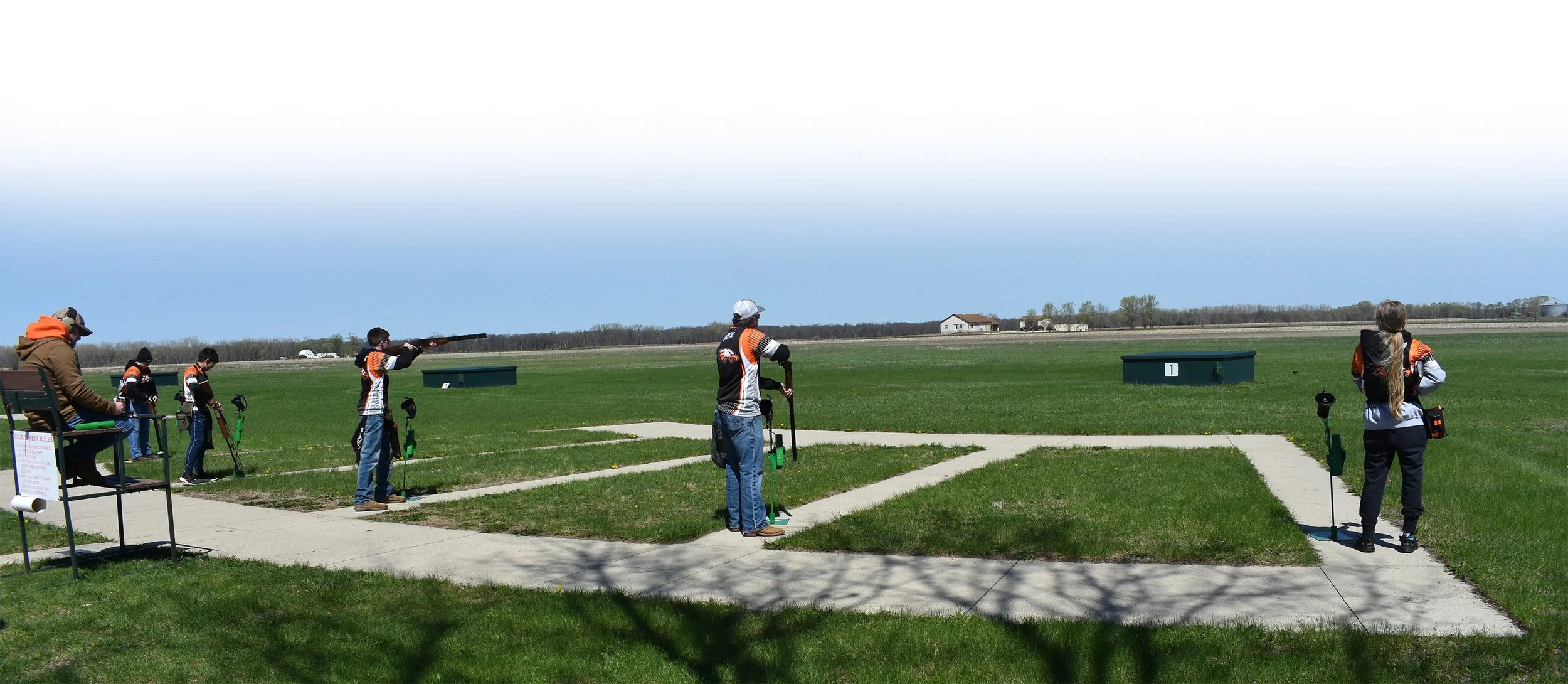 People participating in a shooting sport on a sunny day at a shooting range, aiming rifles at targets downrange, with a grassy field and clear sky in the background.
