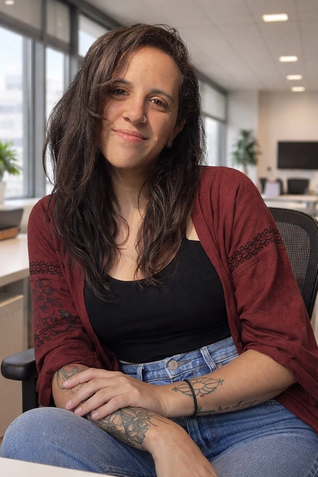 A woman with long, dark wavy hair and a nose piercing, smiling while sitting in an office. She wears a black top, a red cardigan, and blue jeans, with tattoos on her arms. Large windows and office furniture are in the background.