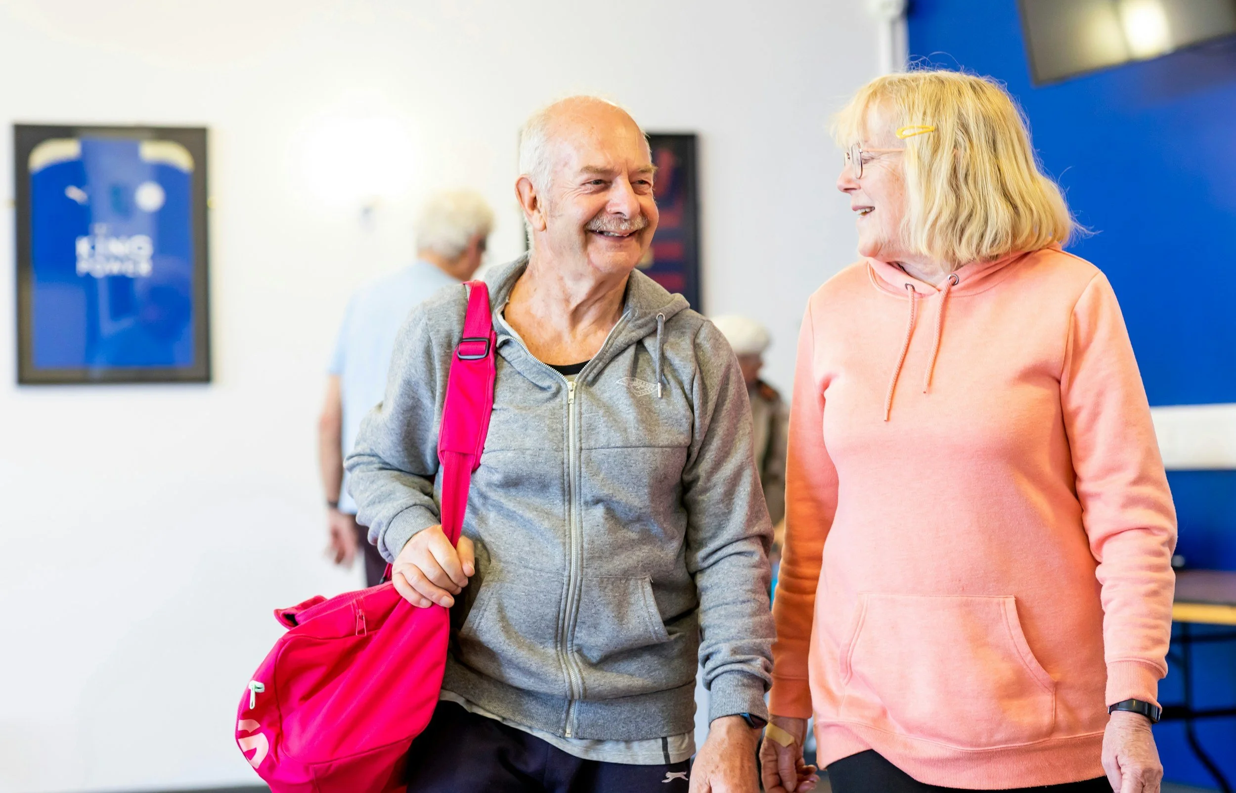 Two elderly women walking and smiling at each other in an indoor setting, with colorful walls and a partially visible basketball jersey on a framed wall in the background.