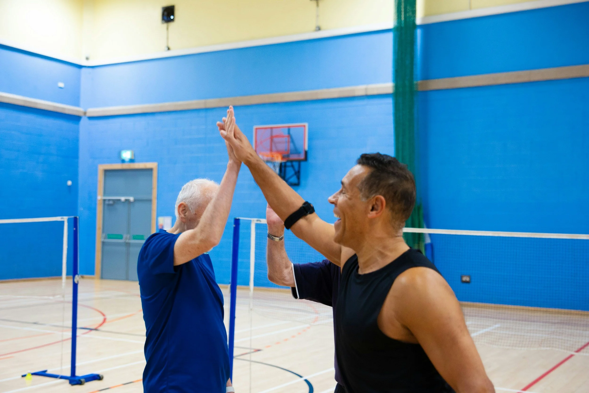 A young man and an elderly man high-fiving in a gymnasium with badminton courts.