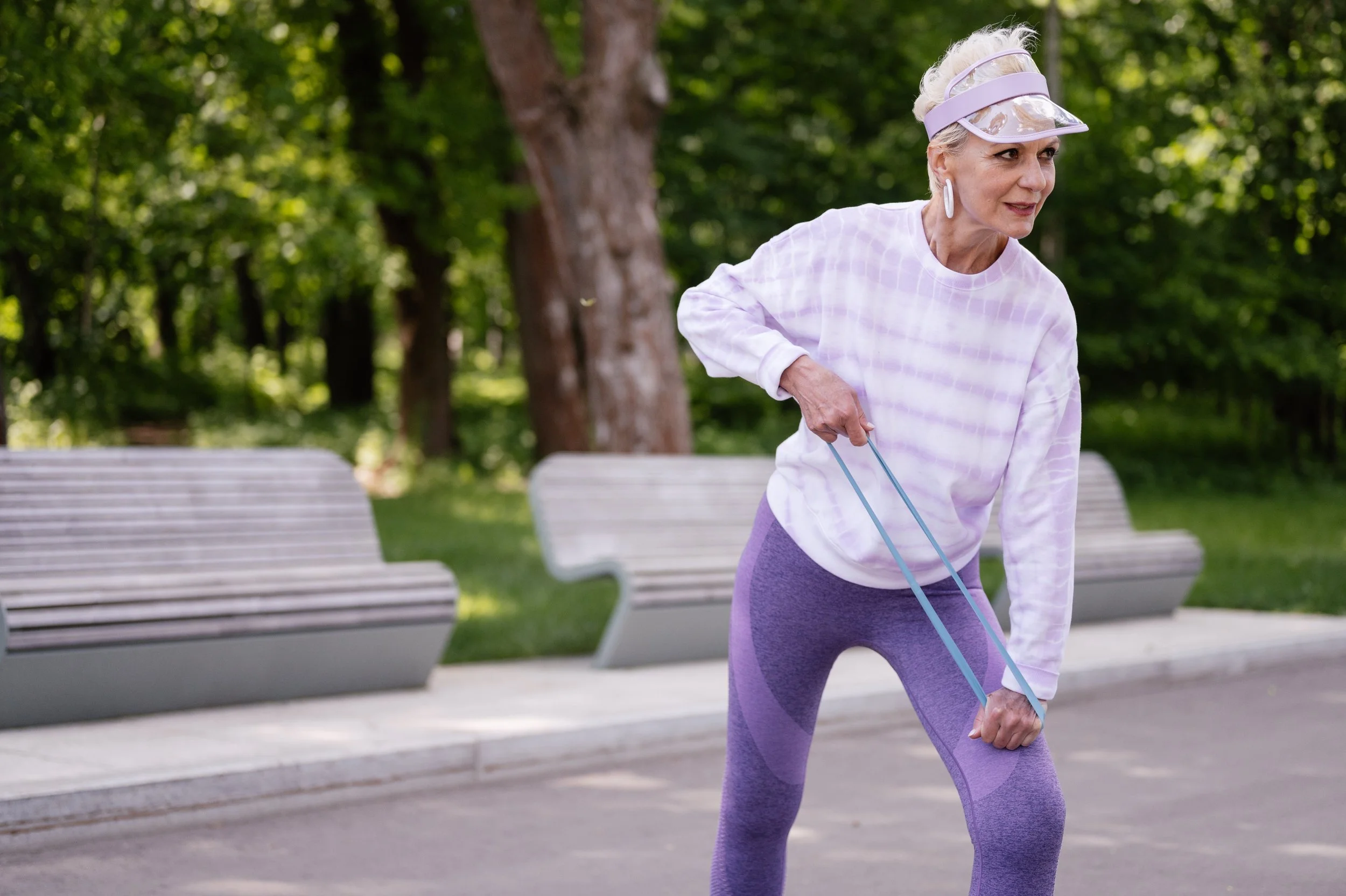 An elderly woman with short gray hair, wearing a purple visor, white sweatshirt, and purple leggings, exercises with resistance bands in a park with green trees and empty benches.
