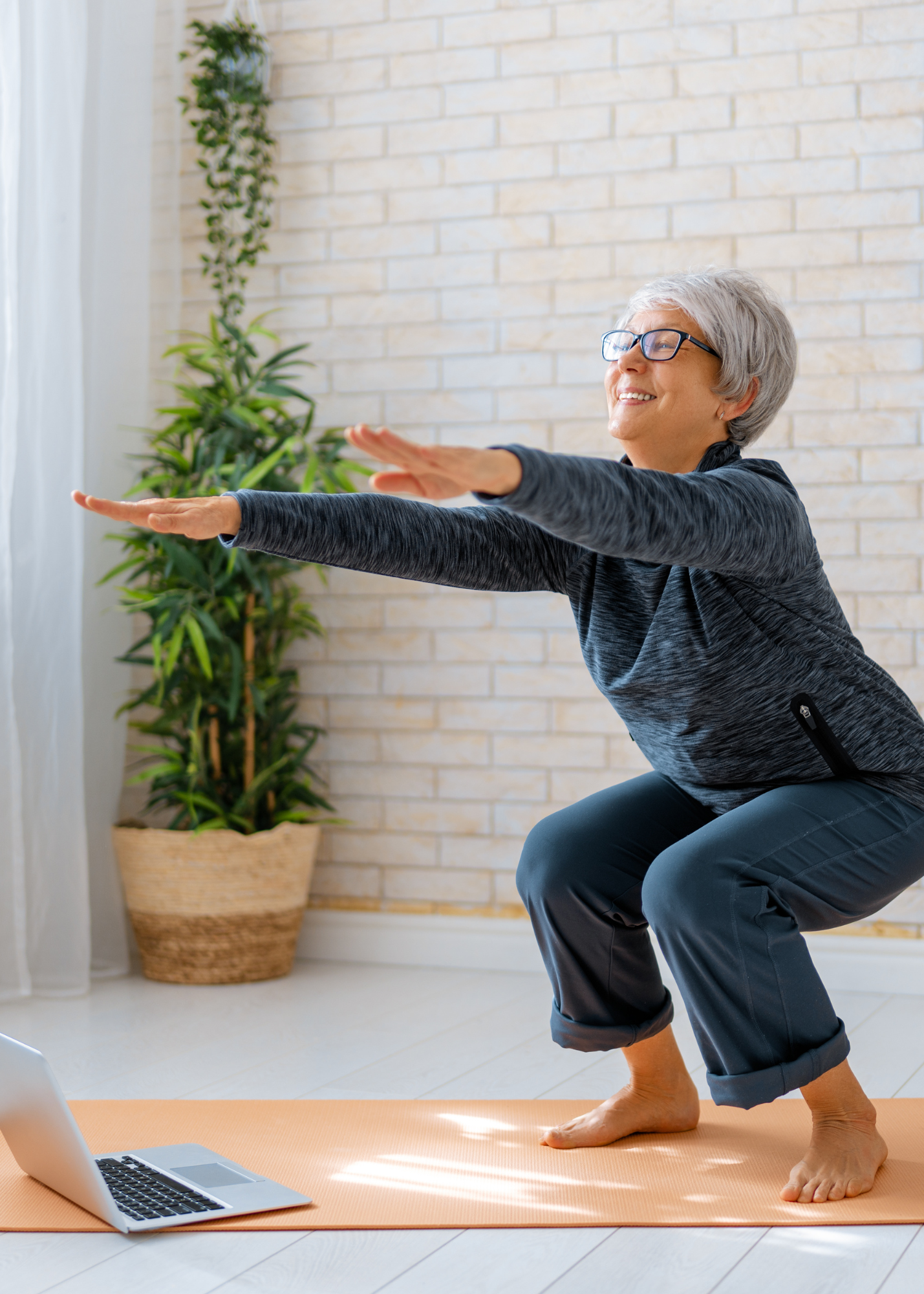 An elderly woman with gray hair and glasses practicing yoga in her living room, doing a squat pose on an orange yoga mat with a laptop in front of her.