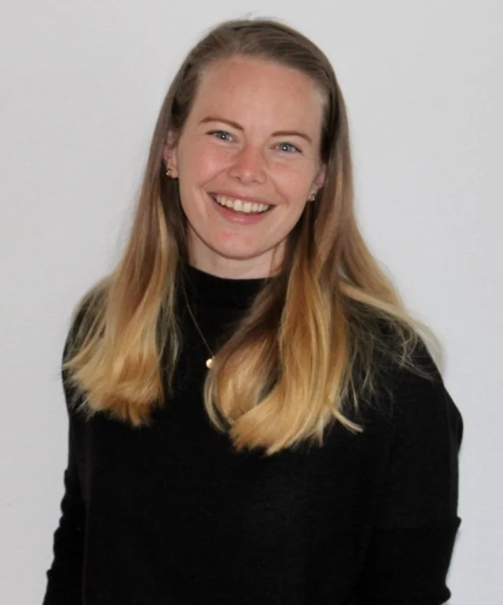 A young woman with long blonde hair, wearing a black top, smiling against a plain white background.