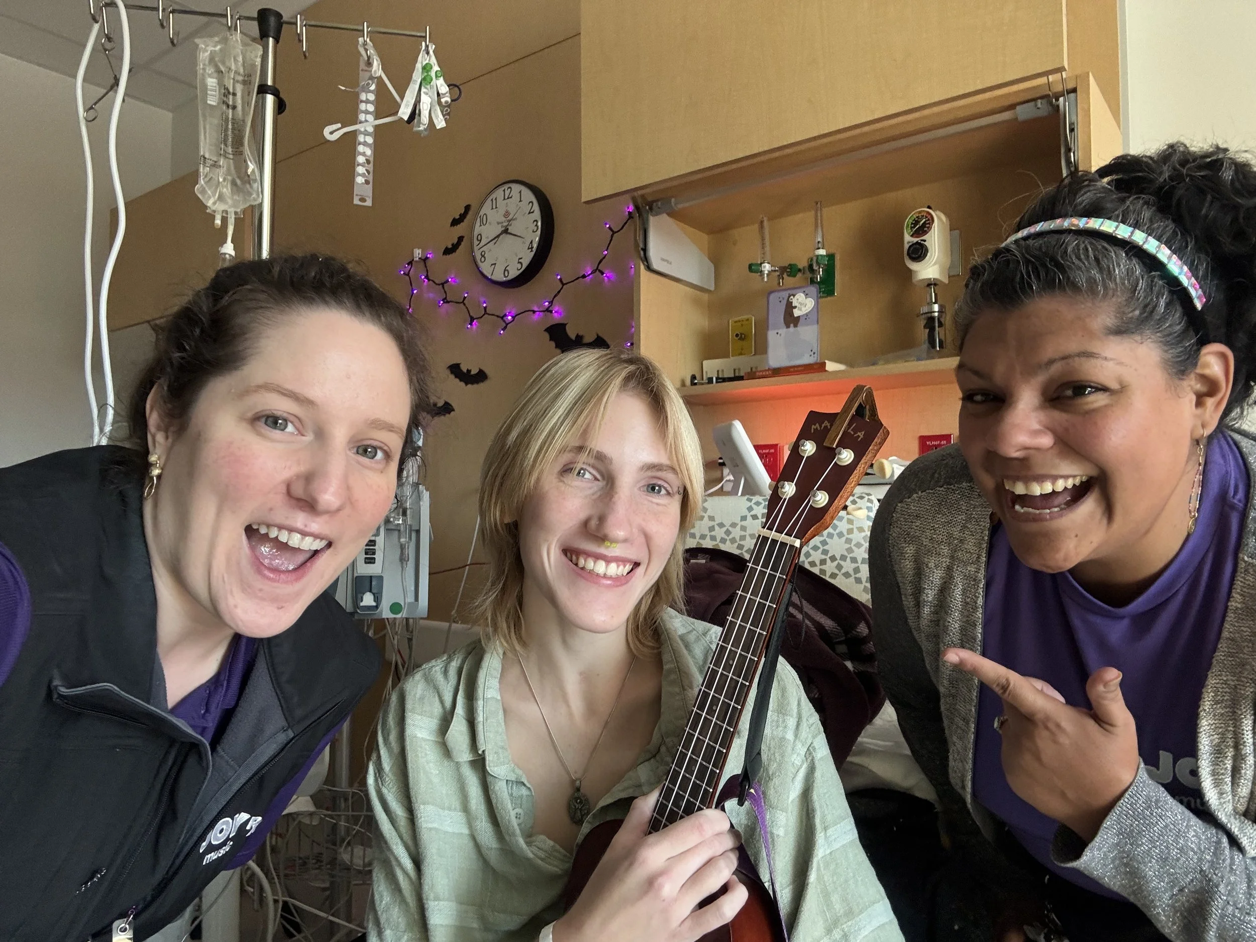 Teenager posing with ukulele in hospital, two JoyRx Music Specialists pointing to the ukulele with big smiles.