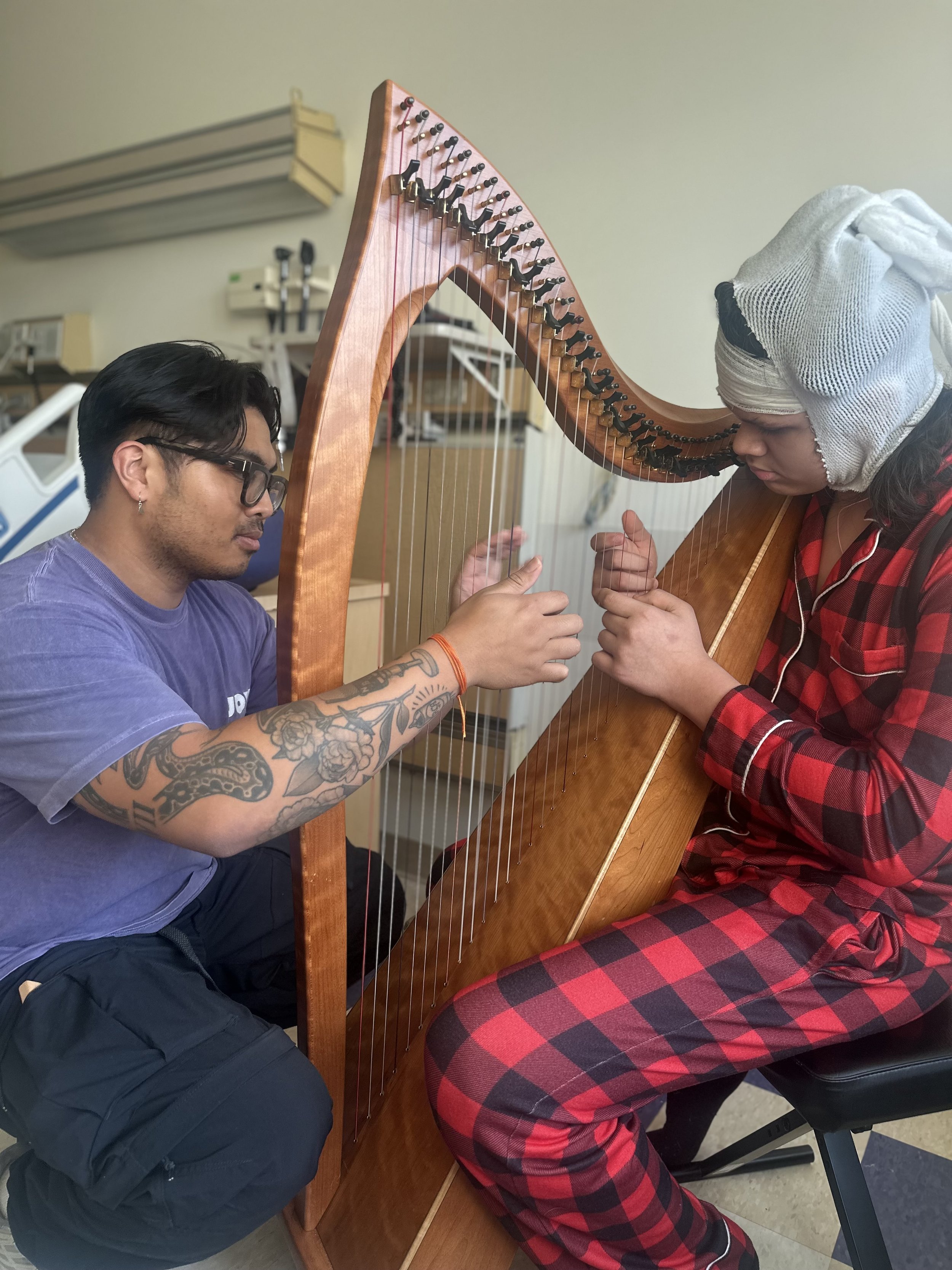 Teen playing the harp at the hospital, being helped by JoyRx Musician in purple shirt and tatoos.