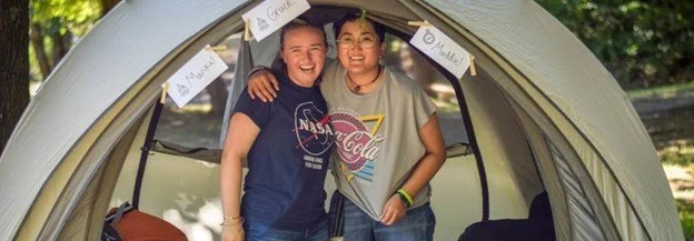 Two teenagers laugh while arm-in-arm in a tent.