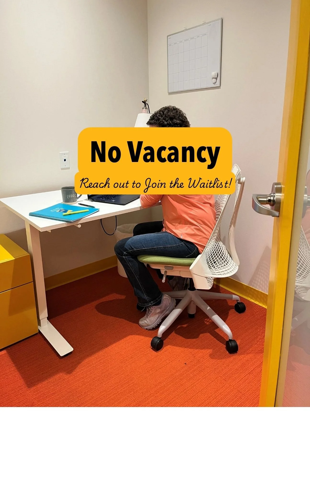 A woman sitting at a white desk working on a laptop in a small, well-lit office with beige walls, a whiteboard, and a yellow door frame.
