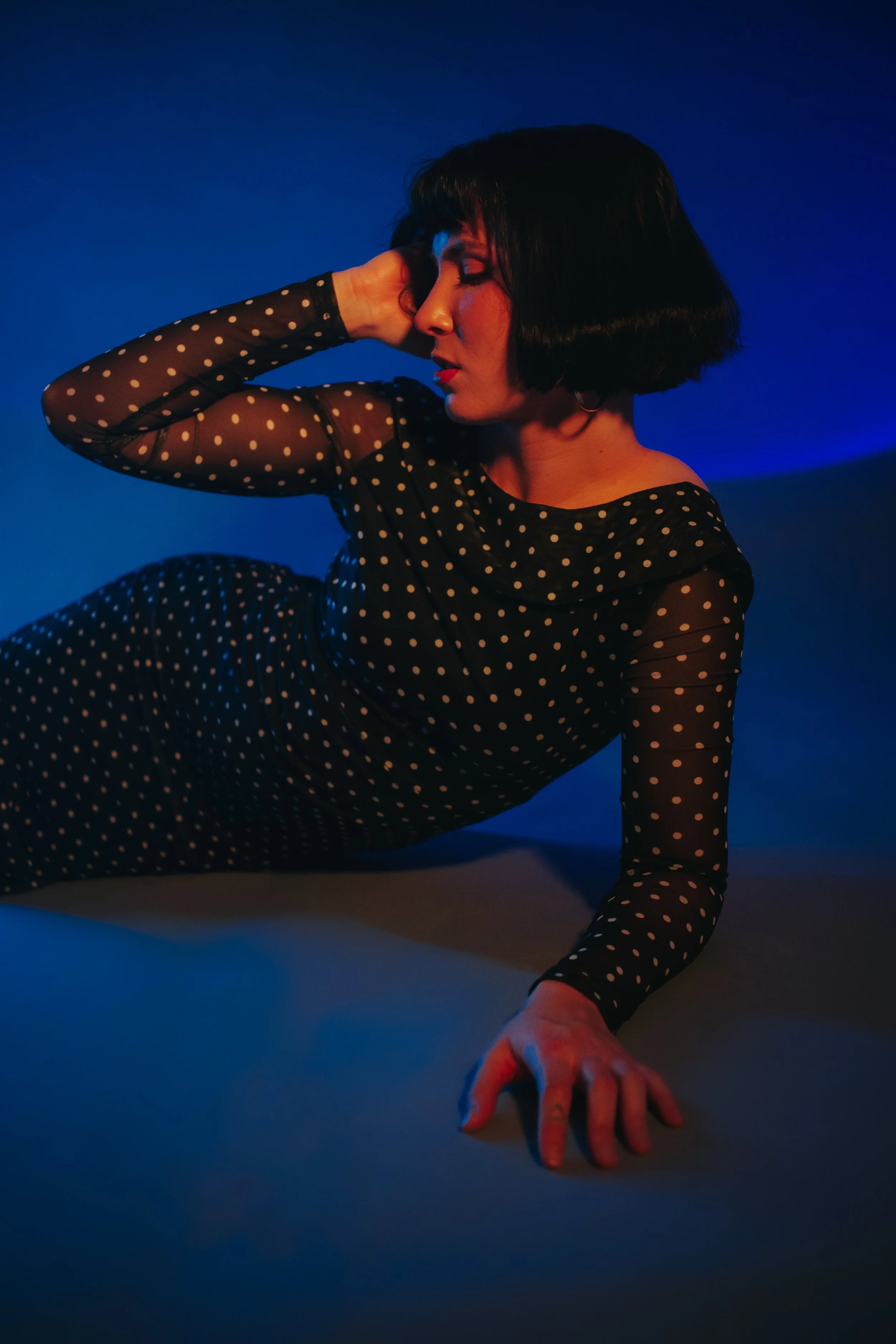 Woman with short black hair in a black polka dot dress posing against a dark blue background.