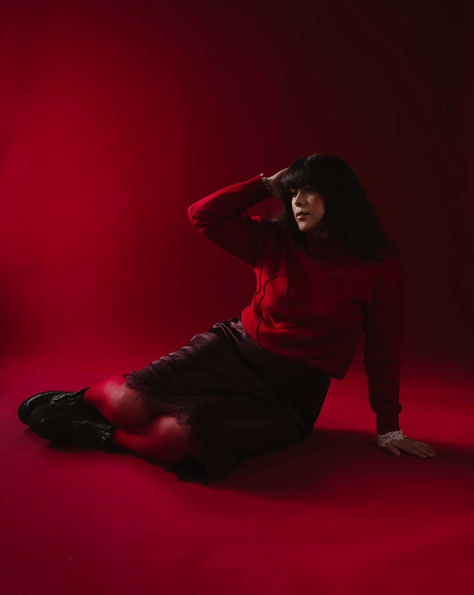 A woman sitting on the floor with one hand touching her head, wearing a red sweater and a black lace skirt, against a red and black background.