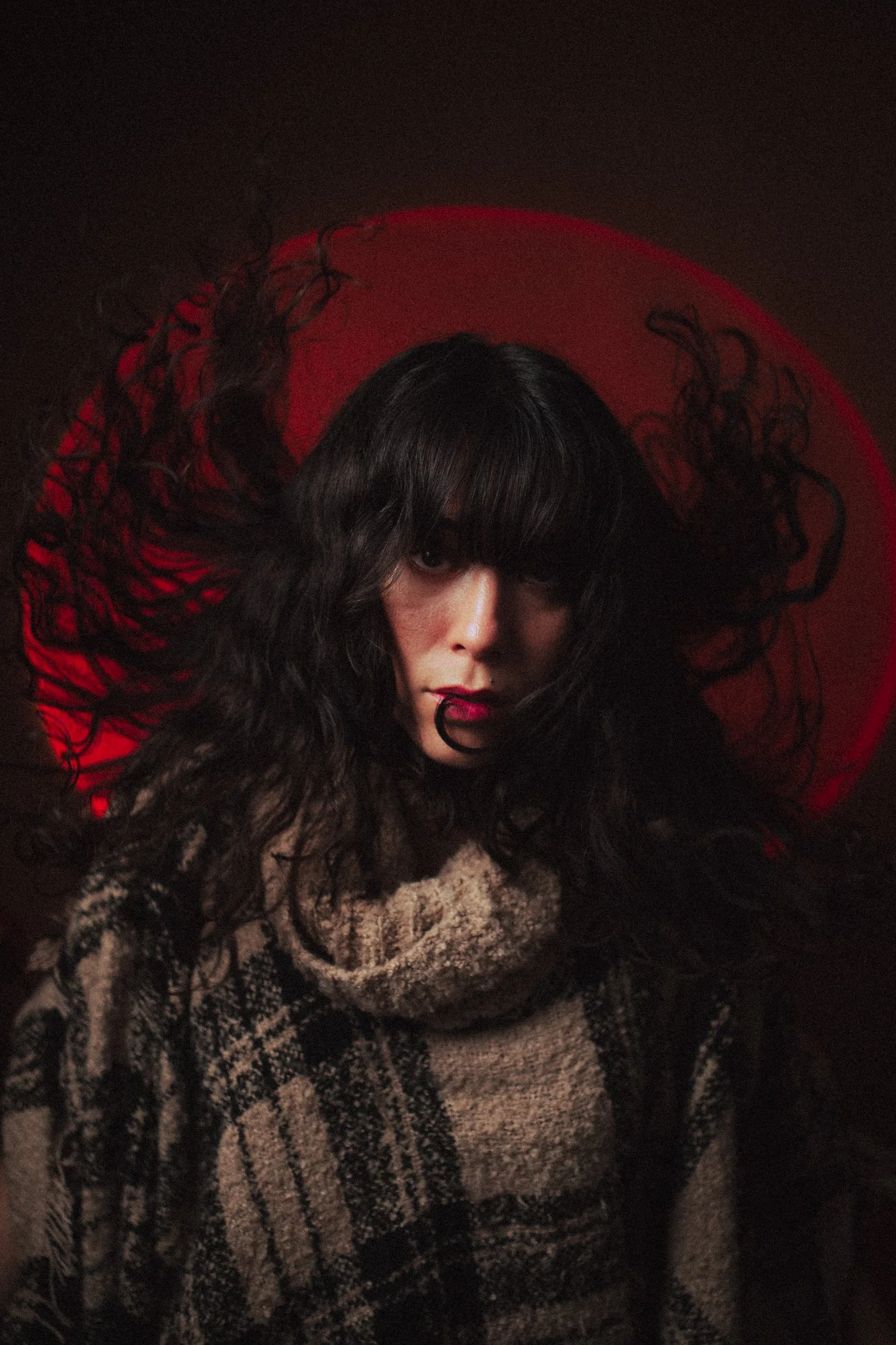 A woman with long, curly dark hair and bangs, wearing a patterned sweater, with a red moon in the background.
