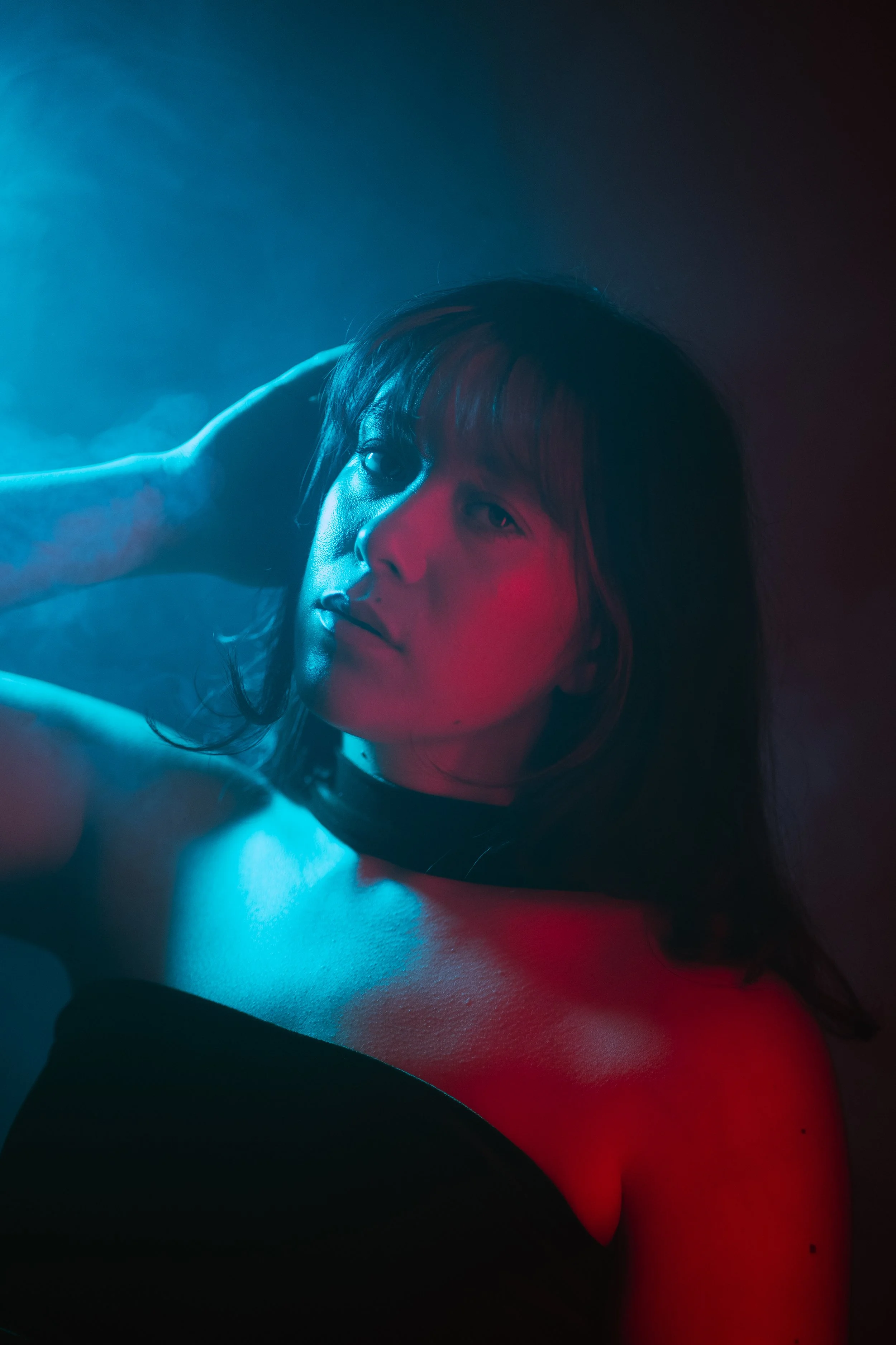 A woman with dark hair and light skin looking at the camera, illuminated with blue and pink lighting with a smoky background.