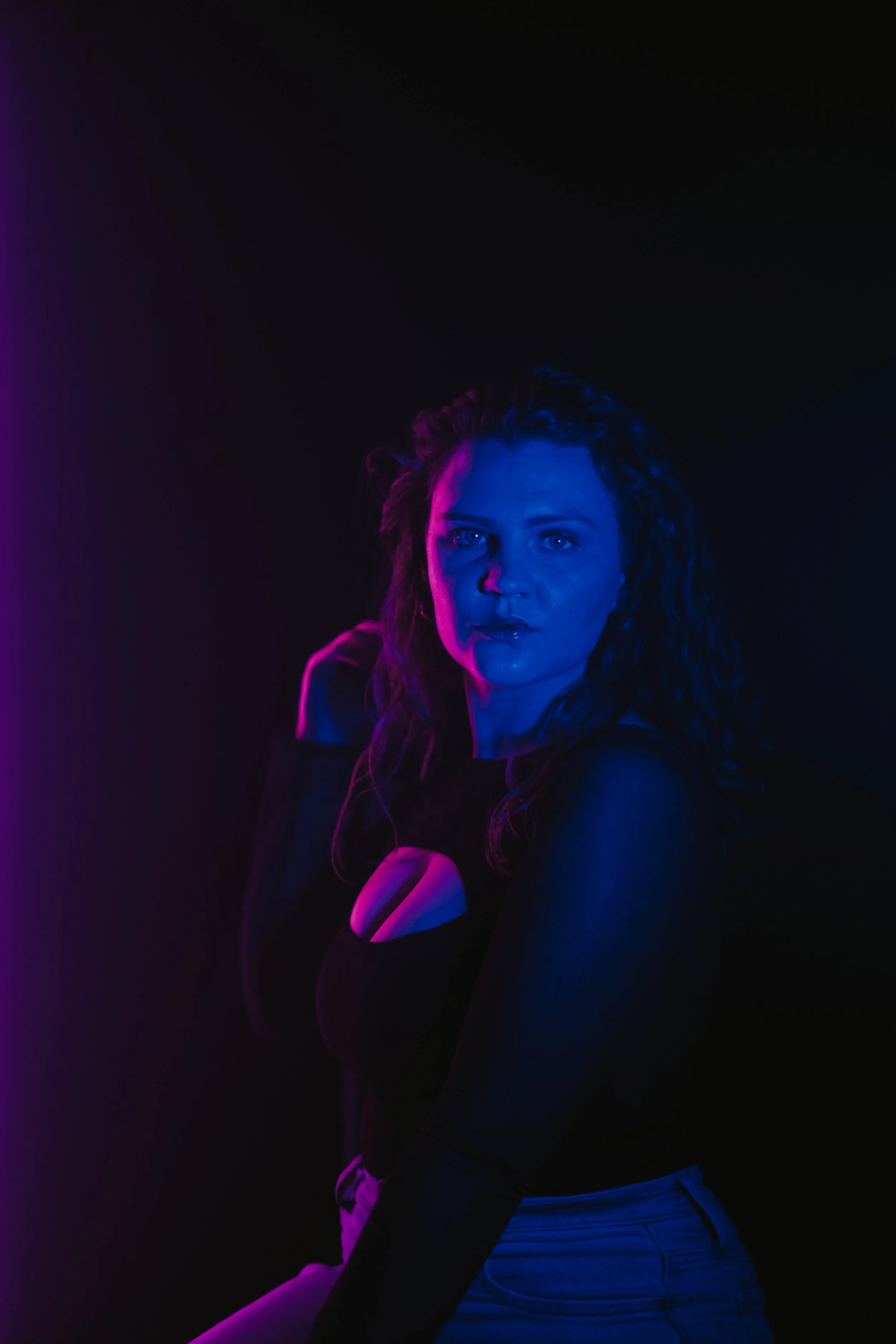 A woman with curly hair illuminated by blue and purple lighting, wearing a black top with a cutout at the chest, against a dark background.