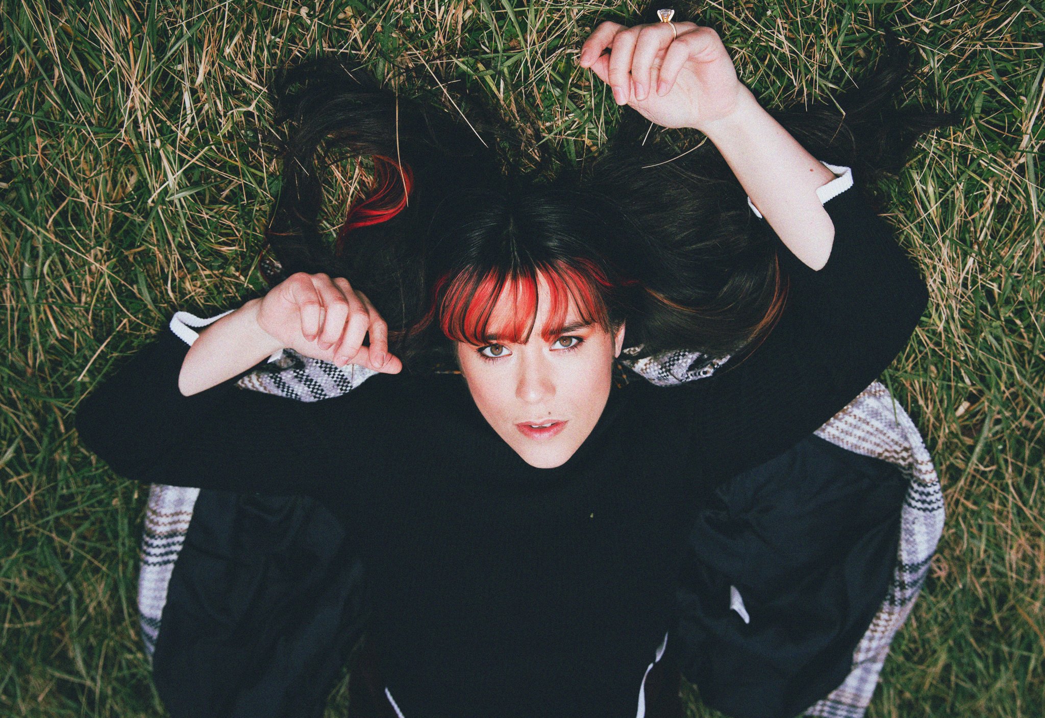 A young woman with dark hair and red streaks, lying on her back in the grass, looking up at the camera.
