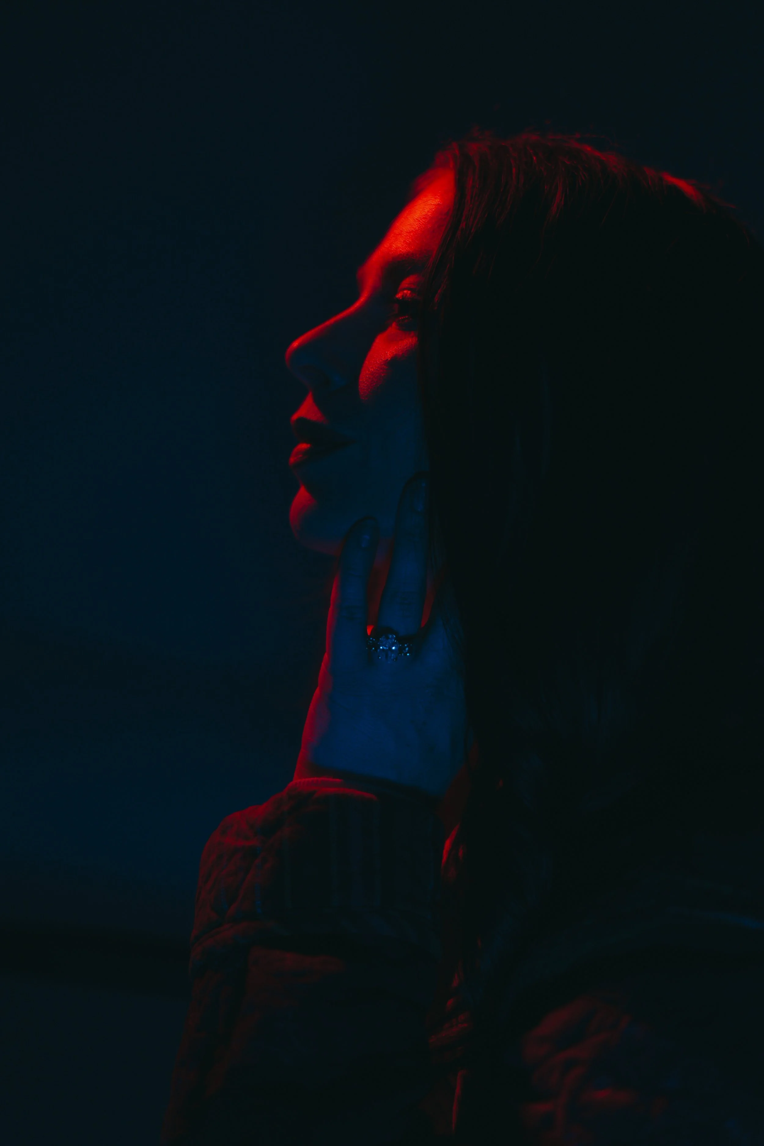 Side profile of a woman’s face and hand with her fingers touching her neck, illuminated by red and blue lighting, dark background