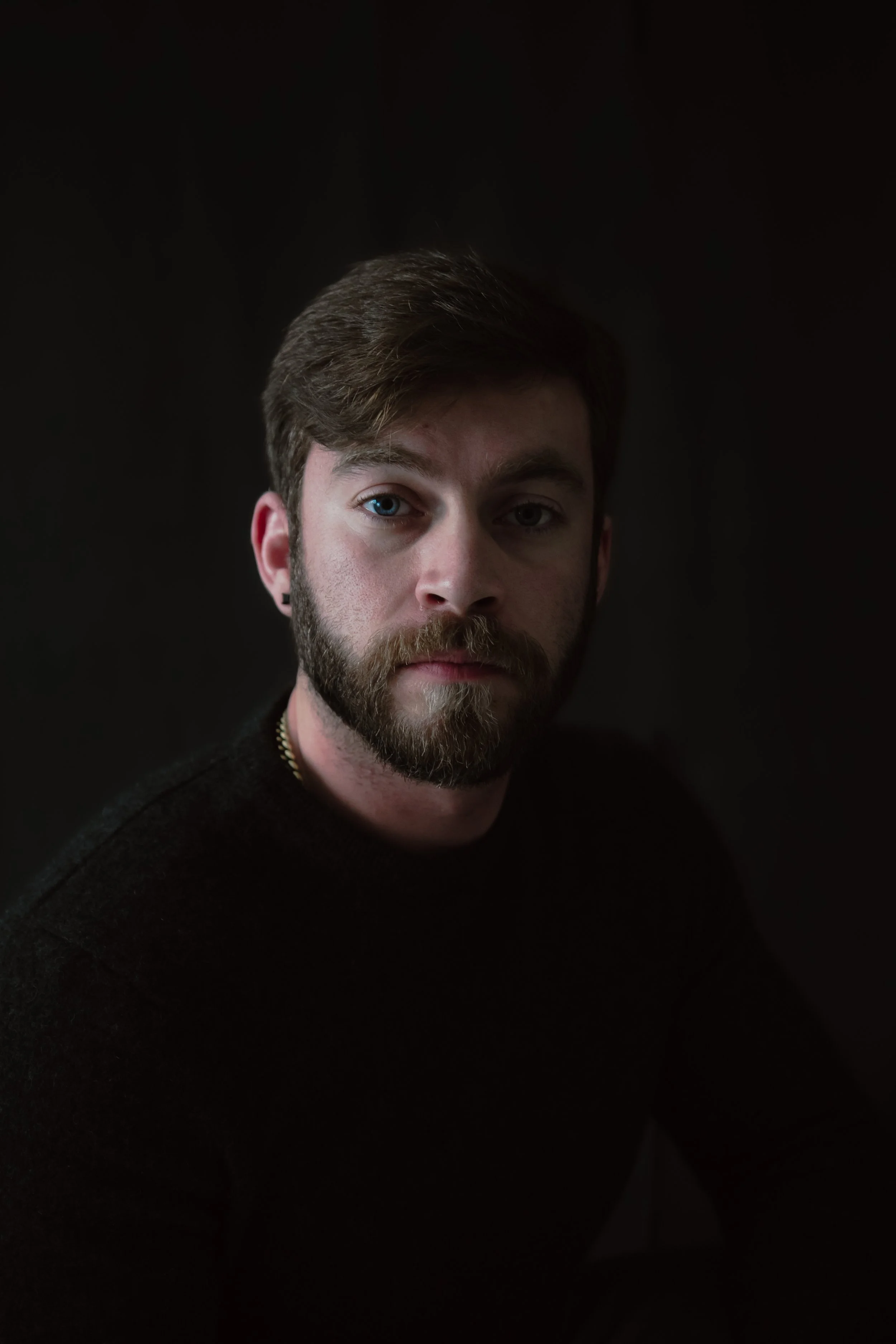 Portrait of a man with brown hair, blue eyes, and a beard, wearing a black shirt, against a dark background.