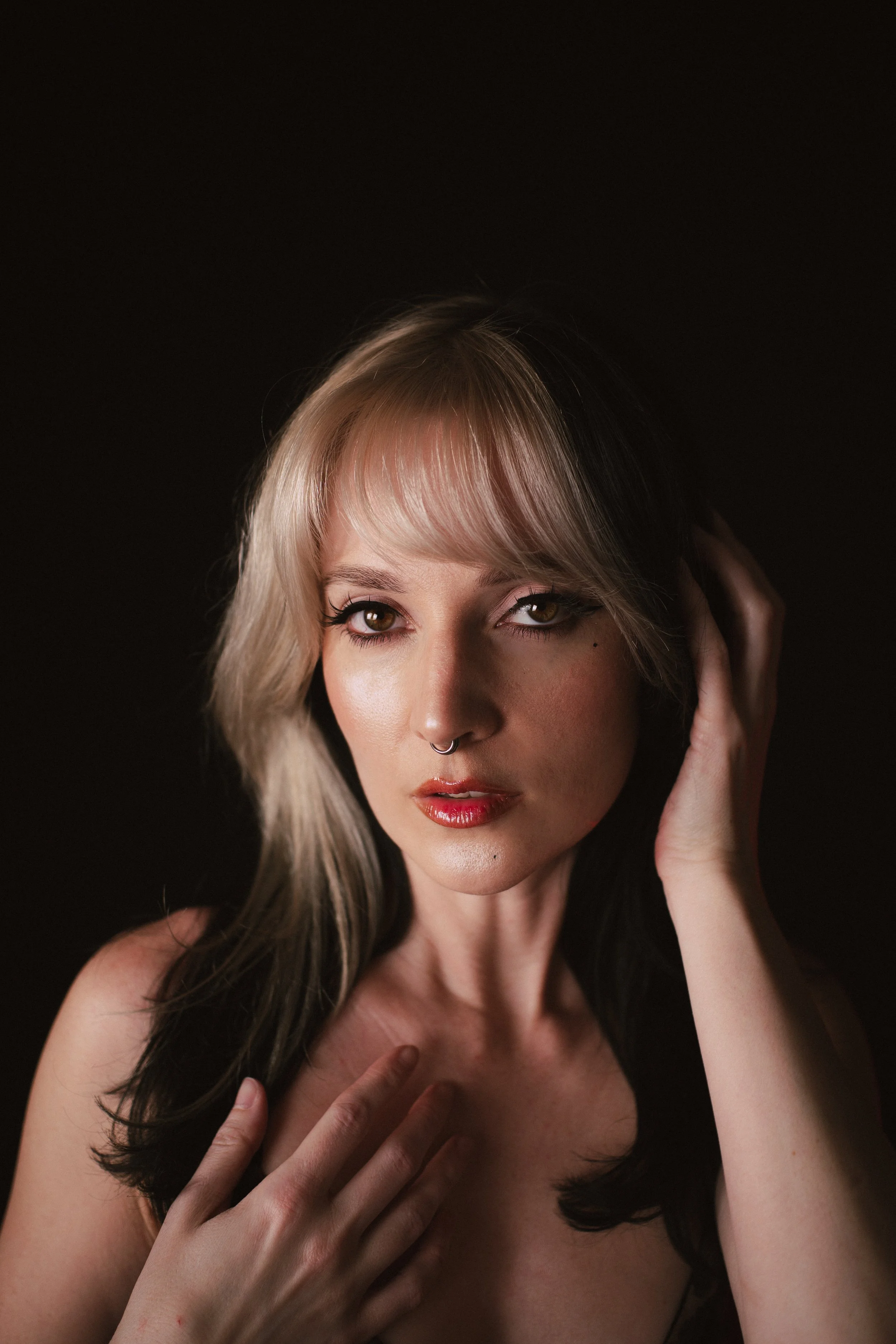 A portrait of a woman with blonde hair, brown eyes, and red lipstick, holding her face with her right hand and her left hand on her chest, against a dark background.