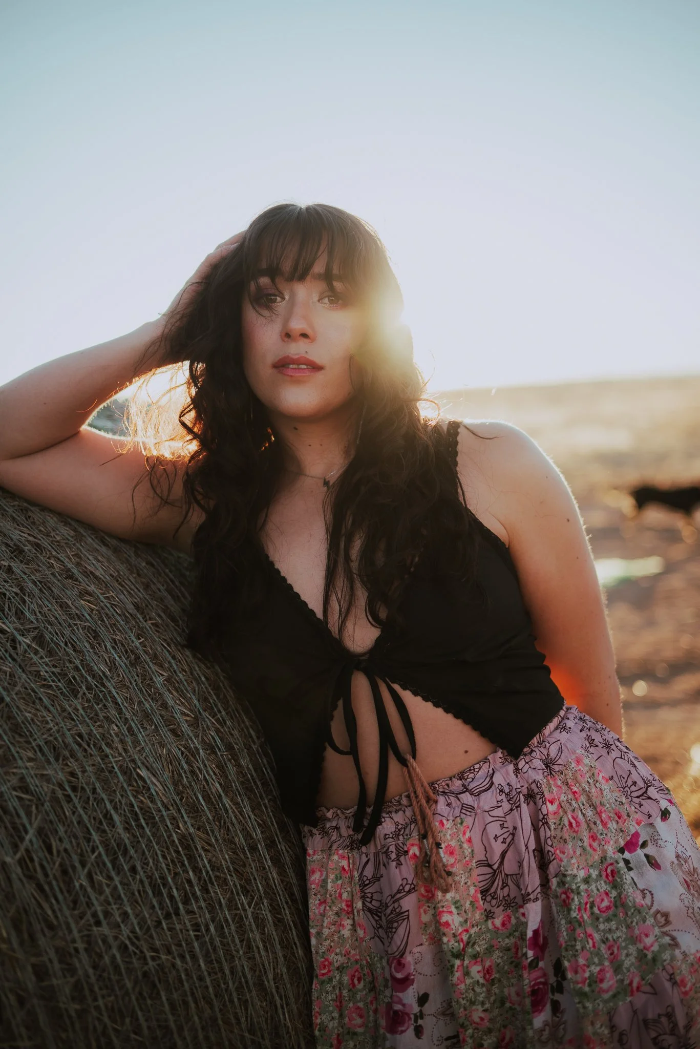 A woman with dark, wavy hair and fair skin, wearing a black top and a colorful, patterned skirt, is leaning against a hay bale outdoors during sunset.