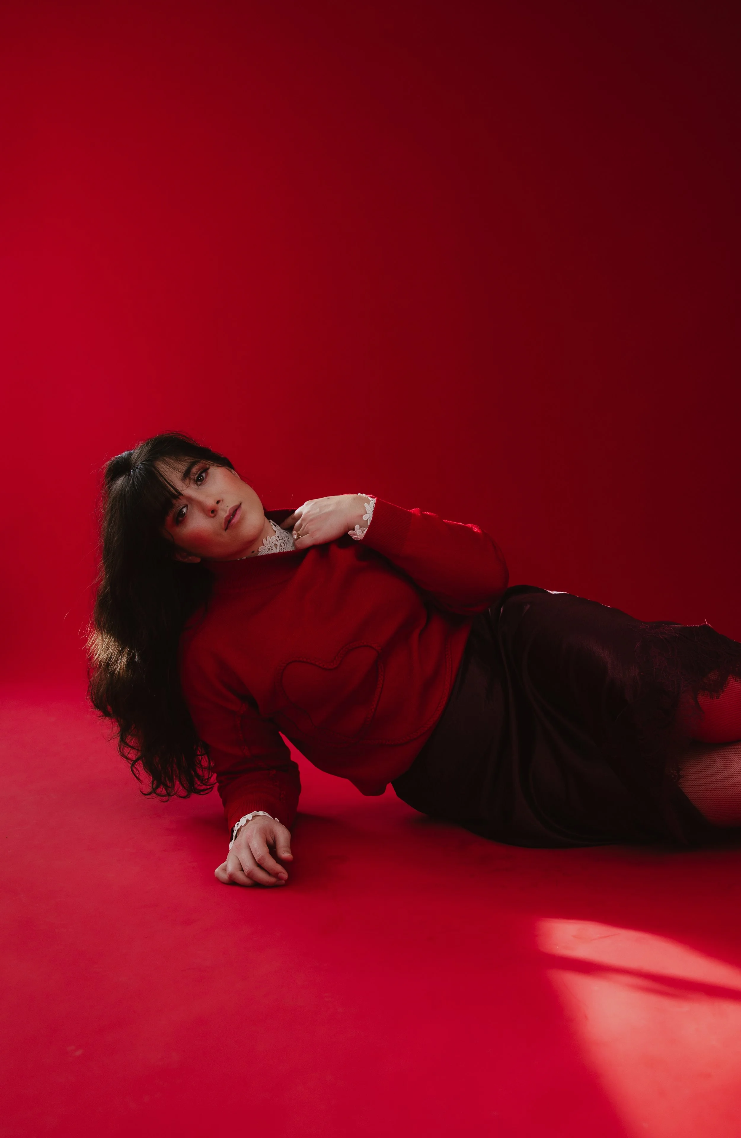 A woman with long dark hair and makeup, wearing a red sweater with white lace details, lying on a red surface against a red background, looking at the camera.