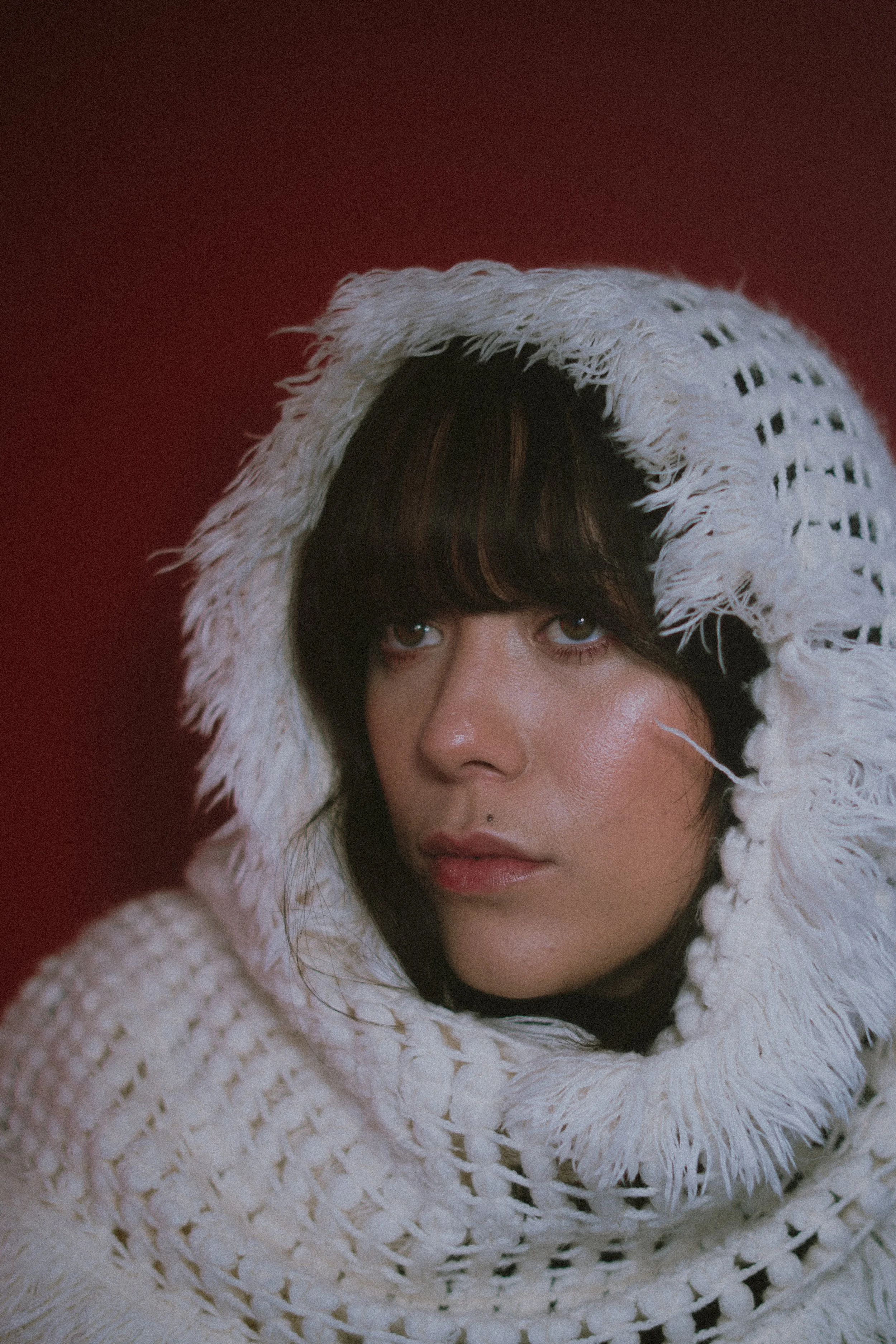 A woman with short dark hair and light skin, wearing a large white fringed knitted hooded garment, against a red background.