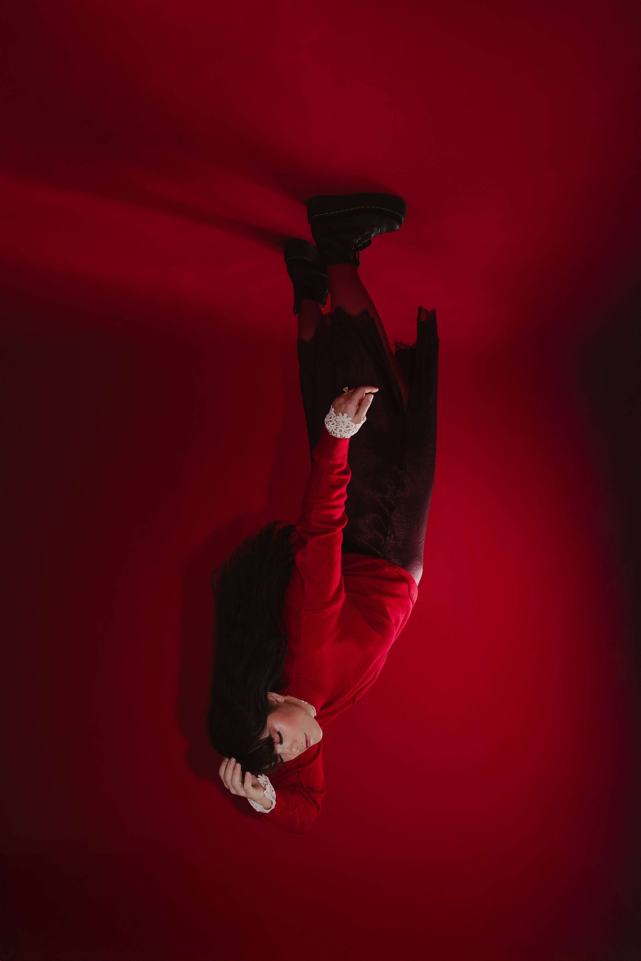 A woman lying on a red surface, wearing a red top, black skirt, and black shoes, with her head resting on her hand and long dark hair.