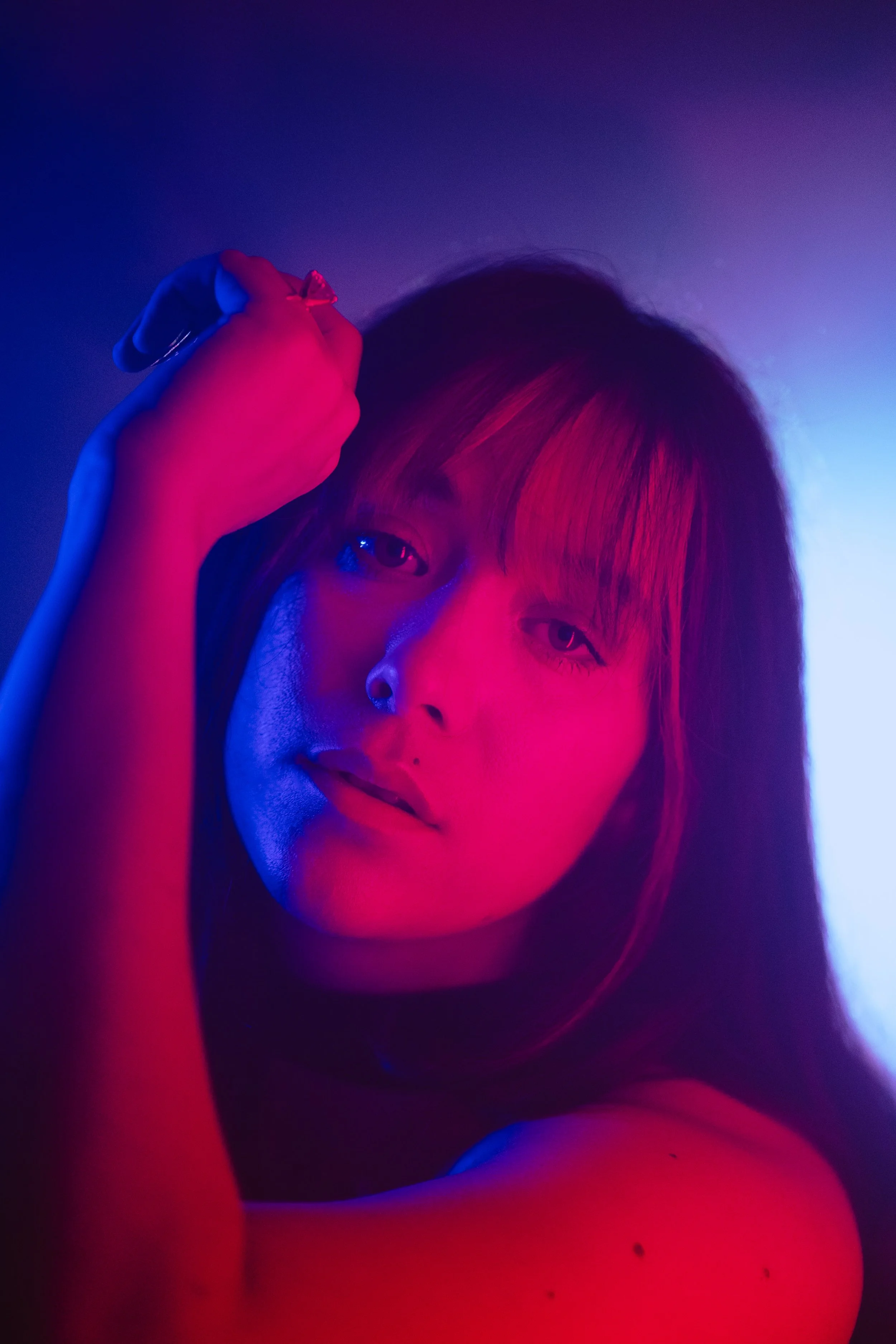 A portrait of a woman with dark hair illuminated by pink and blue neon lighting, looking at the camera with her hand near her head.