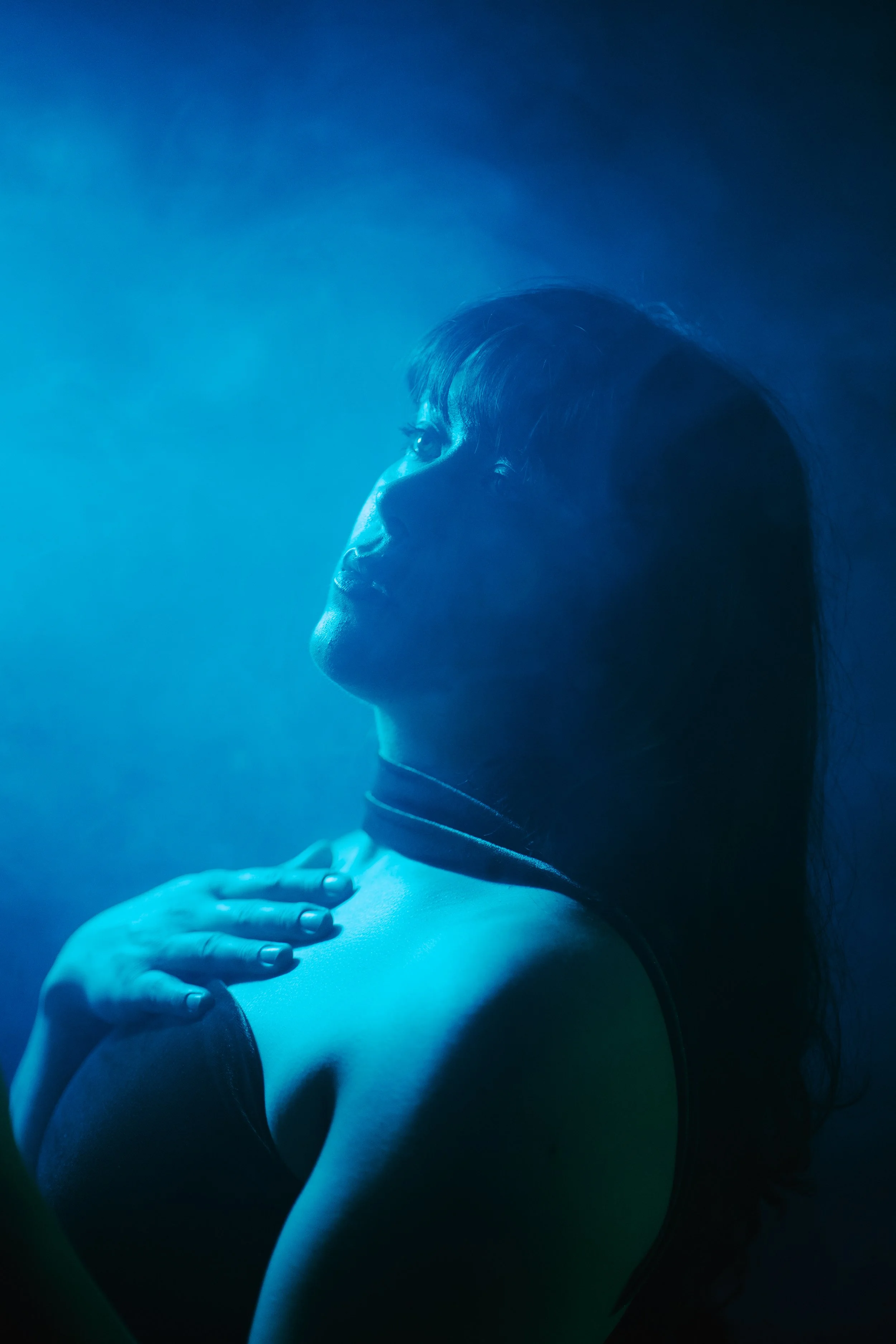 A woman with dark hair and light skin is illuminated by blue light, looking upwards with her hand resting on her shoulder.