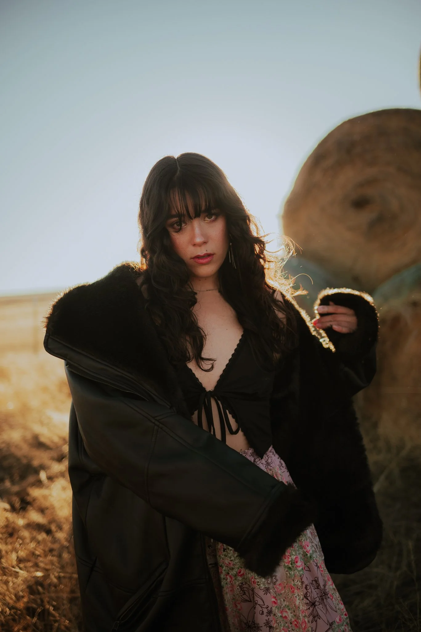 A young woman with dark, curly hair and bangs standing outdoors near logs, illuminated by warm sunlight.
