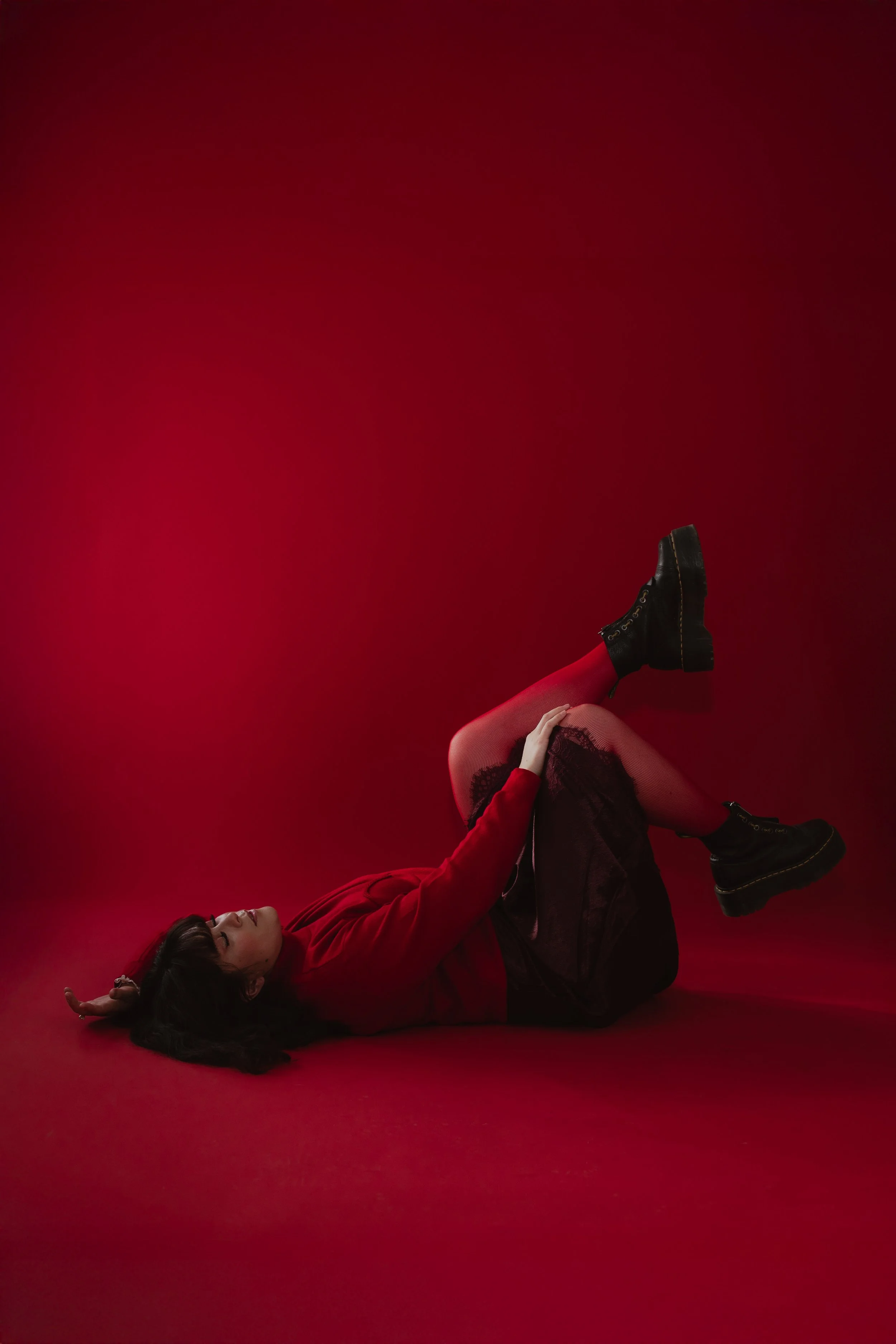 A woman lying on her side on a red background, wearing a red top, a black skirt, red tights, and black platform boots, with one arm resting on her thigh and the other beside her head.