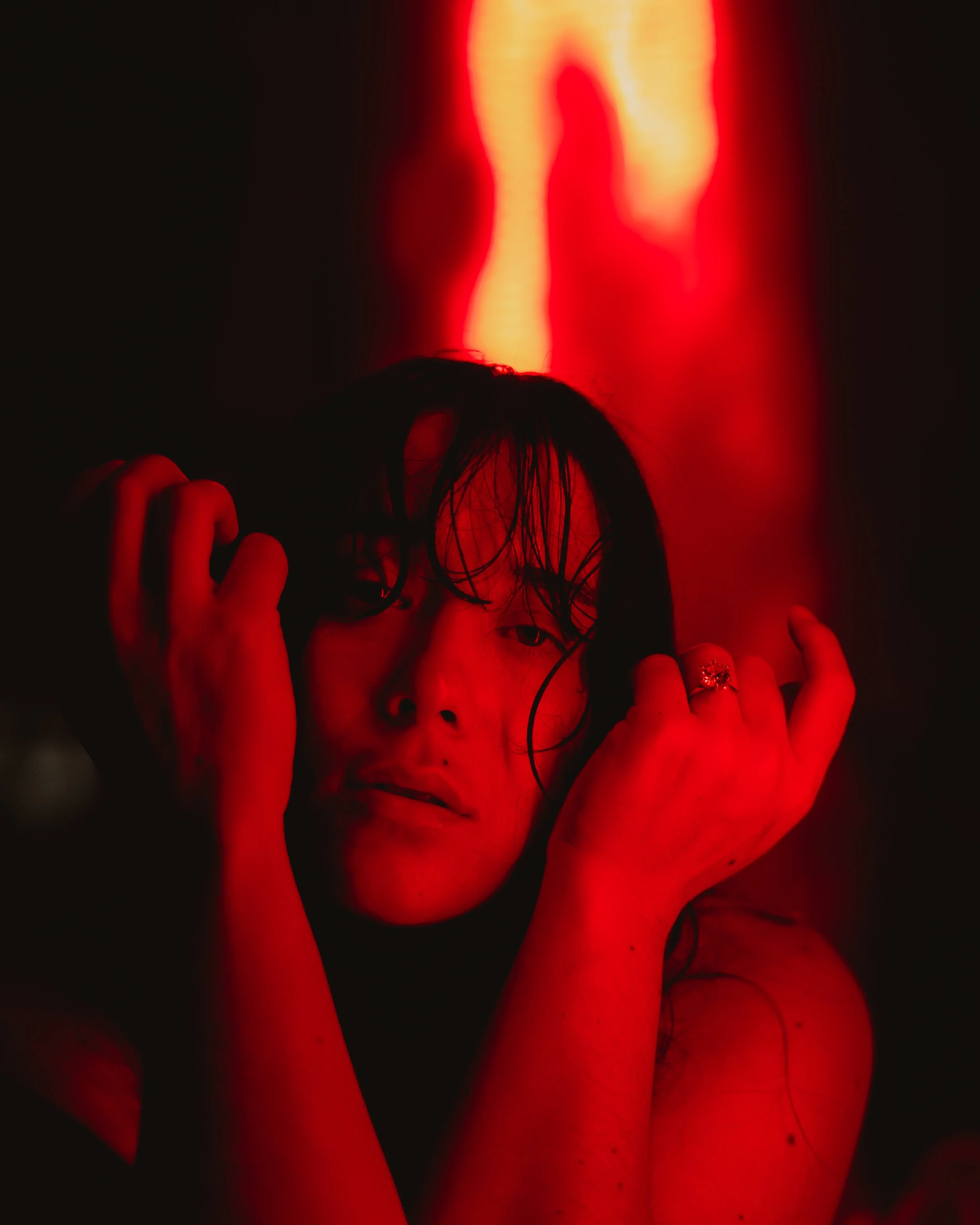 A woman with wet hair posing in front of red lights, holding her head with both hands.