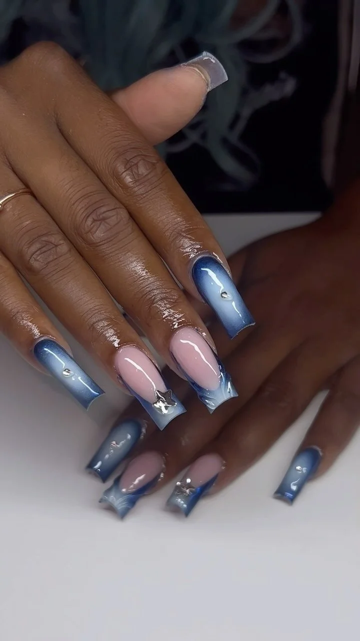 Close-up of a hand with blue and pink ombré acrylic nails decorated with small rhinestones and silver star charms.