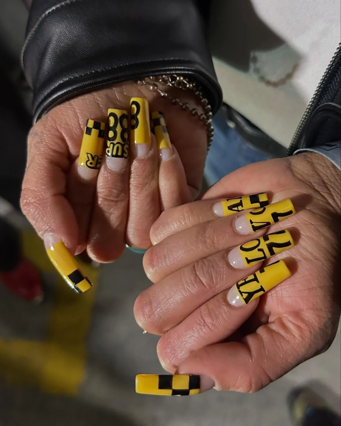 Close-up of hands with yellow and black checkered nails, holding a chain and wearing a black leather jacket.