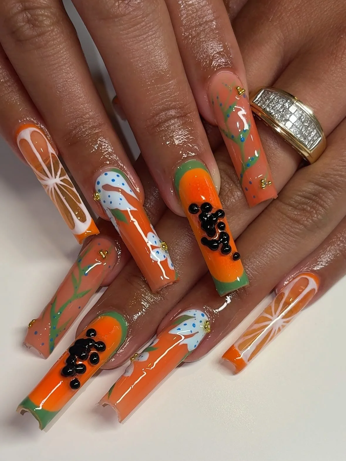Nail art with orange, green, white, blue, and black designs, including flowers, abstract patterns, and small black beads, on long square-shaped nails.