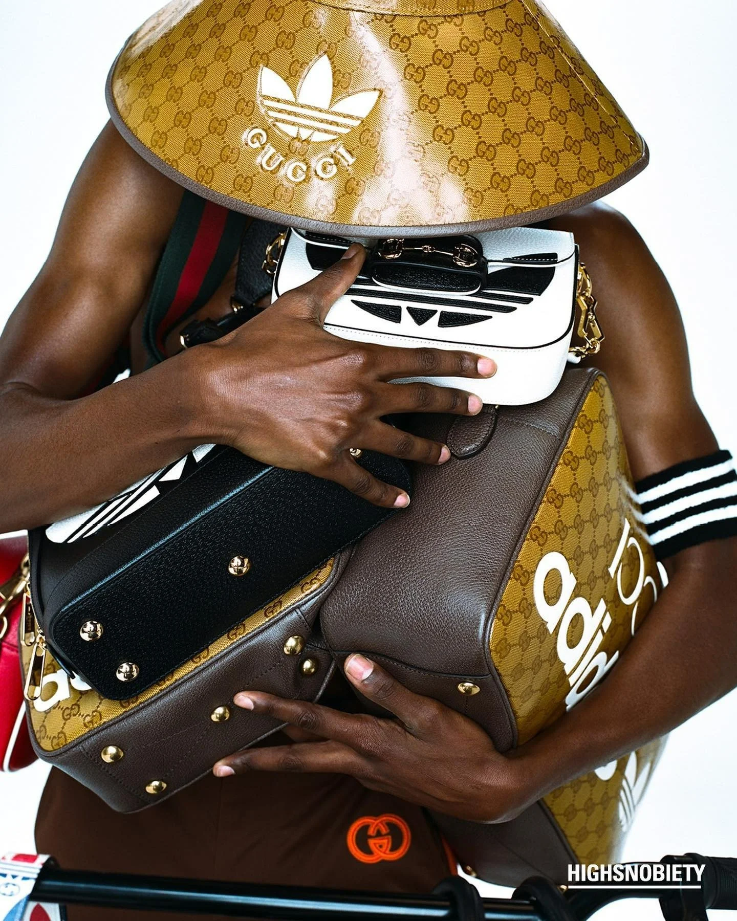 Person holding multiple designer bags and wearing a Gucci hat with the Adidas logo, with a Gucci belt visible at the waist.