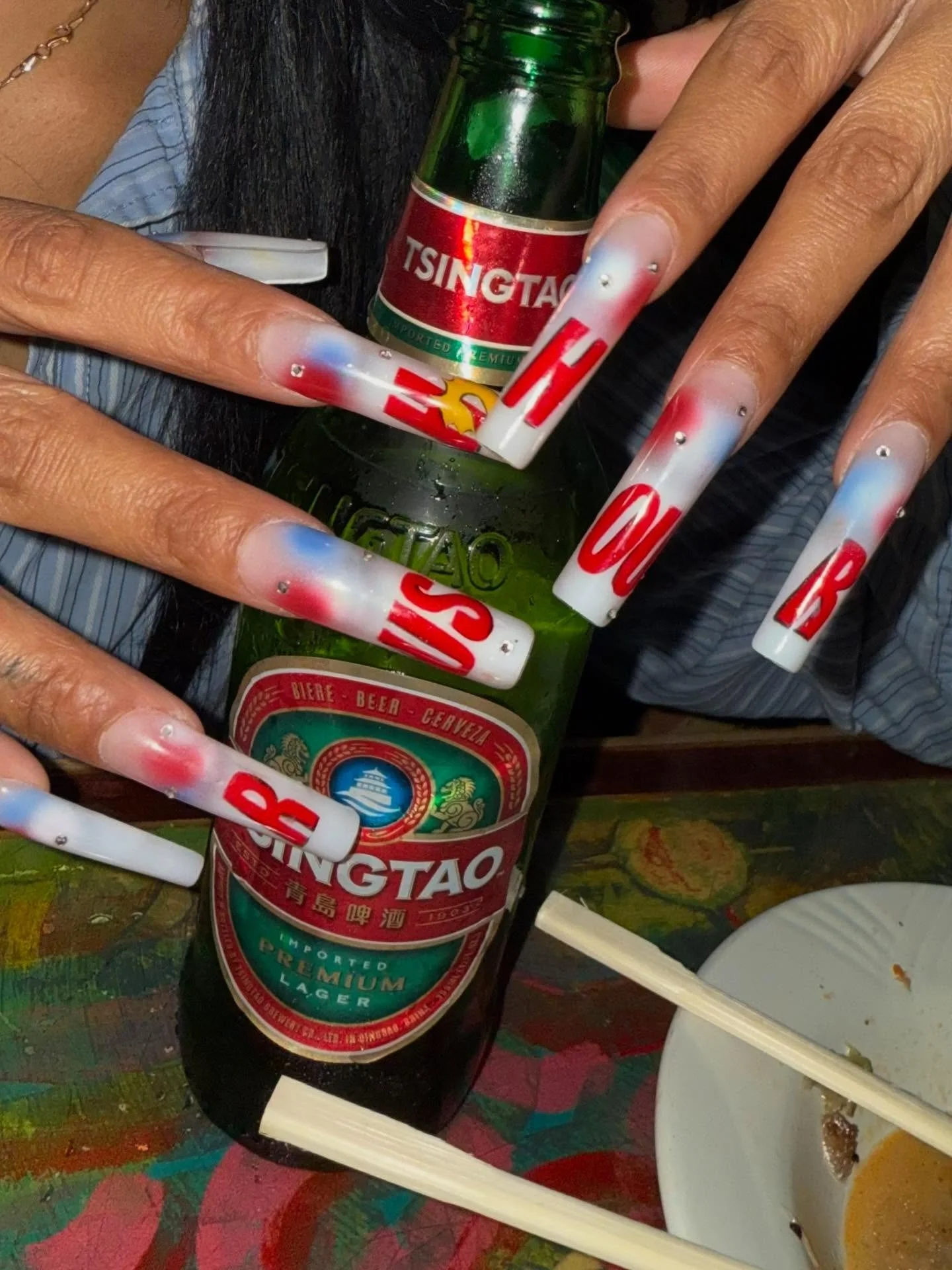 A person with long, decorated nails holding a green bottle of Tsingtao beer with chopsticks beside a white dish on a colorful table.