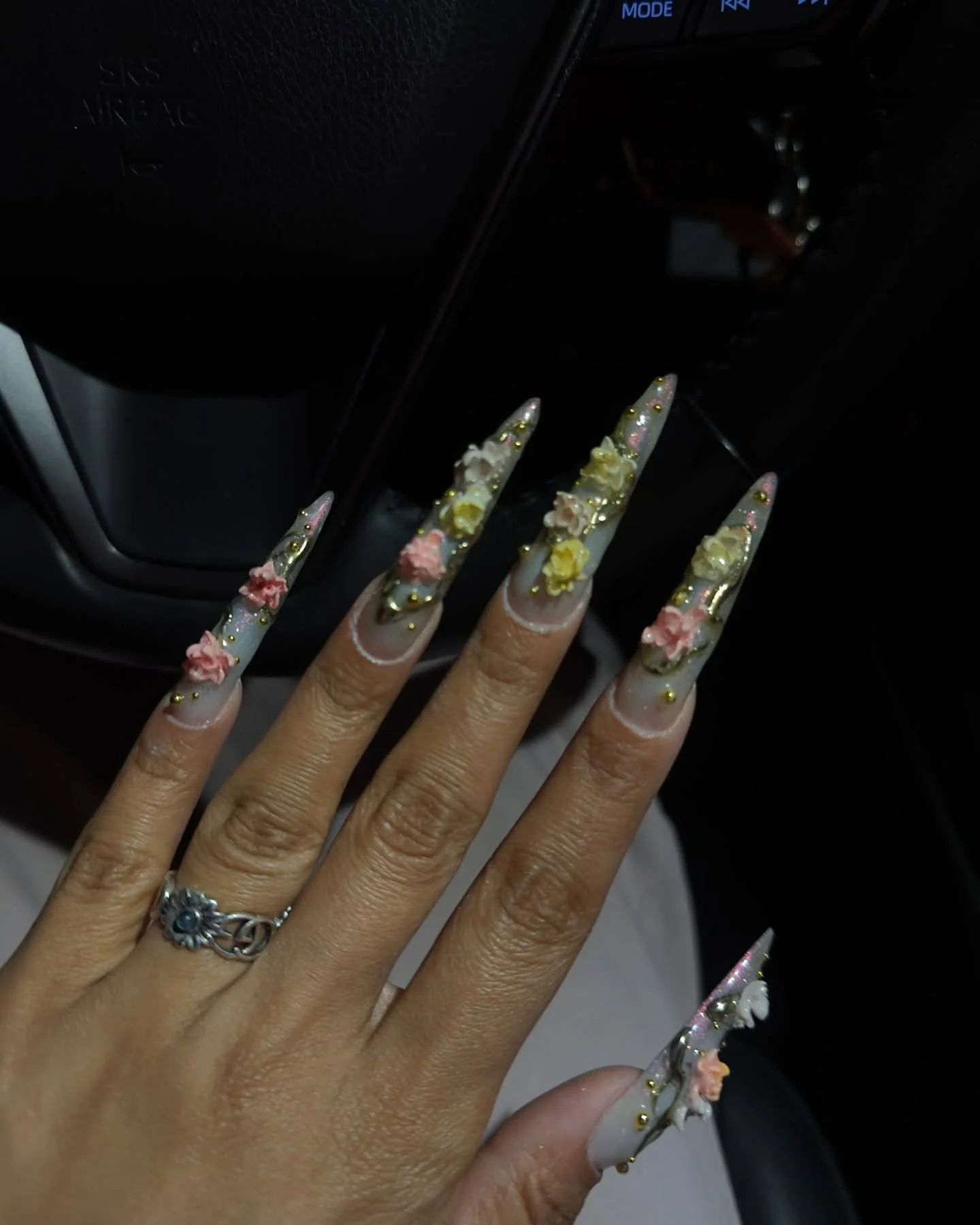 Close-up of a hand with long, decorated nails featuring 3D flower designs and gold accents, inside a vehicle.