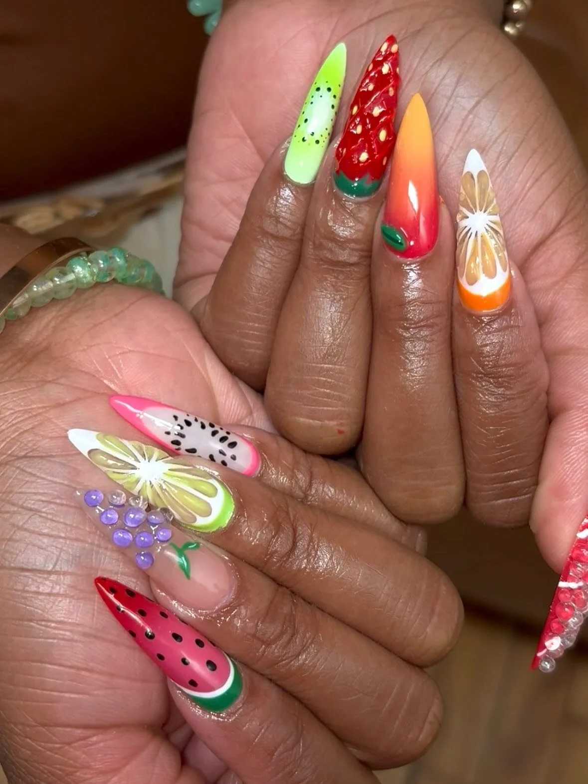 Close-up of hands with colorful, fruit-themed nail art featuring strawberries, watermelon, lemon, grapes, and dragon fruit designs.
