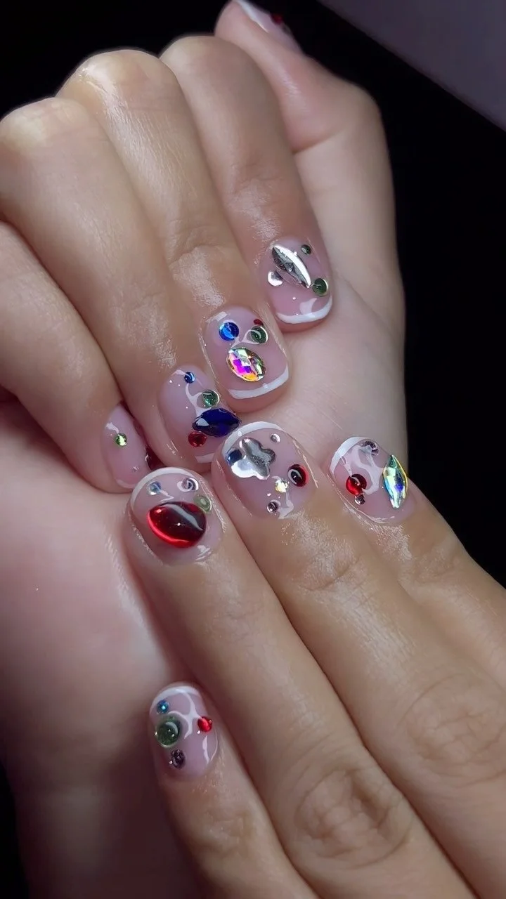 Close-up of a hand with decorated fingernails featuring colorful rhinestones, holographic accents, and intricate designs.