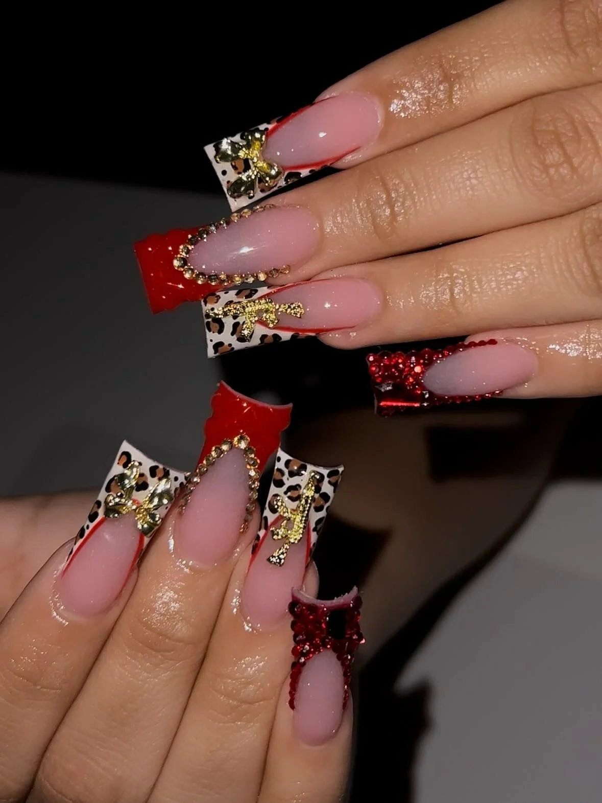 Close-up of hands with elaborately decorated long acrylic nails featuring red, black, and white colors, leopard print patterns, gold embellishments, pearls, and small decorative bows.