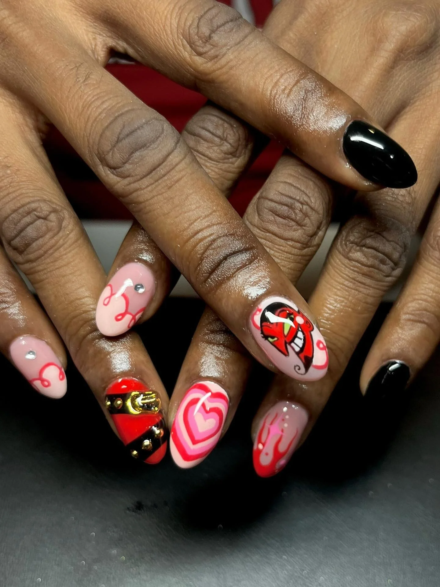 Close-up of manicured nails with pink, red, black, and white designs including hearts, flames, and a cartoon character, some with rhinestones and metallic accents.