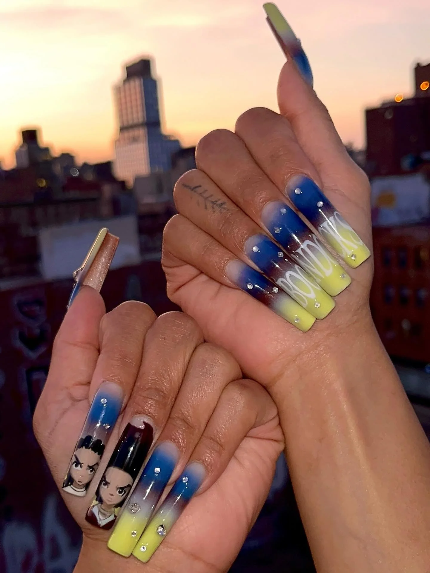 Close-up of a person's hands with colorful, decorated long acrylic nails, set against a city skyline at sunset.