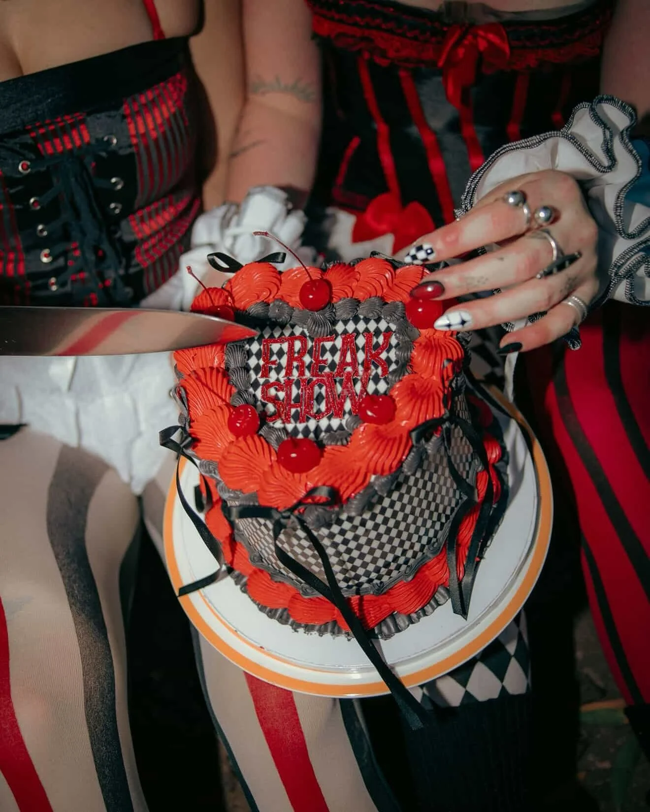 A person is cutting a heart-shaped cake decorated with red and black frosting, cherries, and the words 'FREAK SHOW' written on top. The person has painted nails, rings, and is wearing ruffled sleeves and striped clothing.
