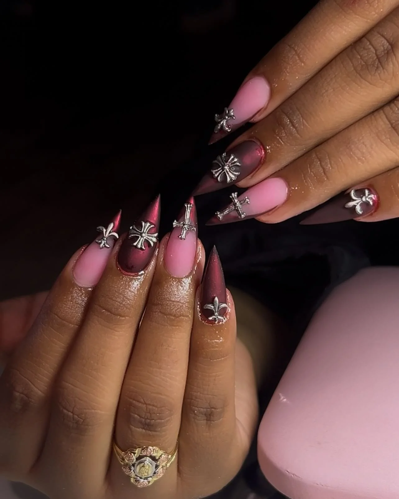 Close-up of hands with elaborately decorated long, pointed nails in pink and burgundy, adorned with silver cross and fleur-de-lis nail charms, and a gold ring with a large clear gemstone.