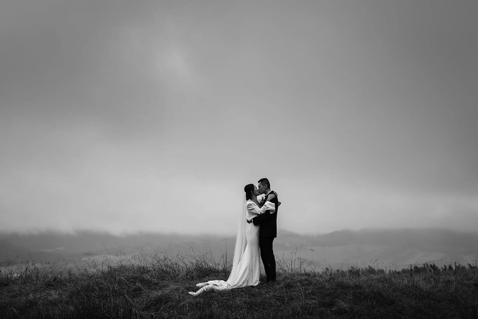 A black-and-white photo of a couple in wedding attire sharing a kiss outdoors, with a cloudy sky and grassy field in the background.