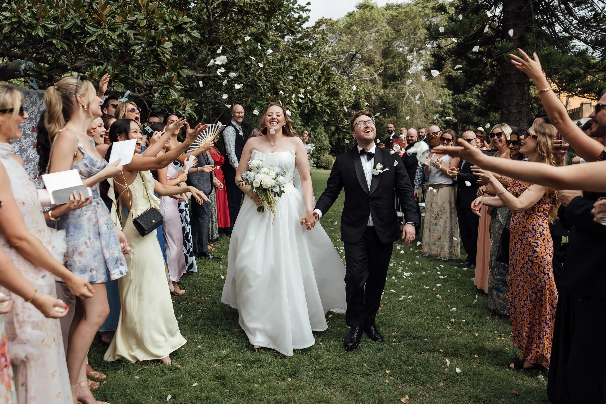 A bride and groom walking hand in hand through a crowd of guests during an outdoor wedding celebration at Vaucluse House Estate, with confetti falling around them, surrounded by trees and greenery.