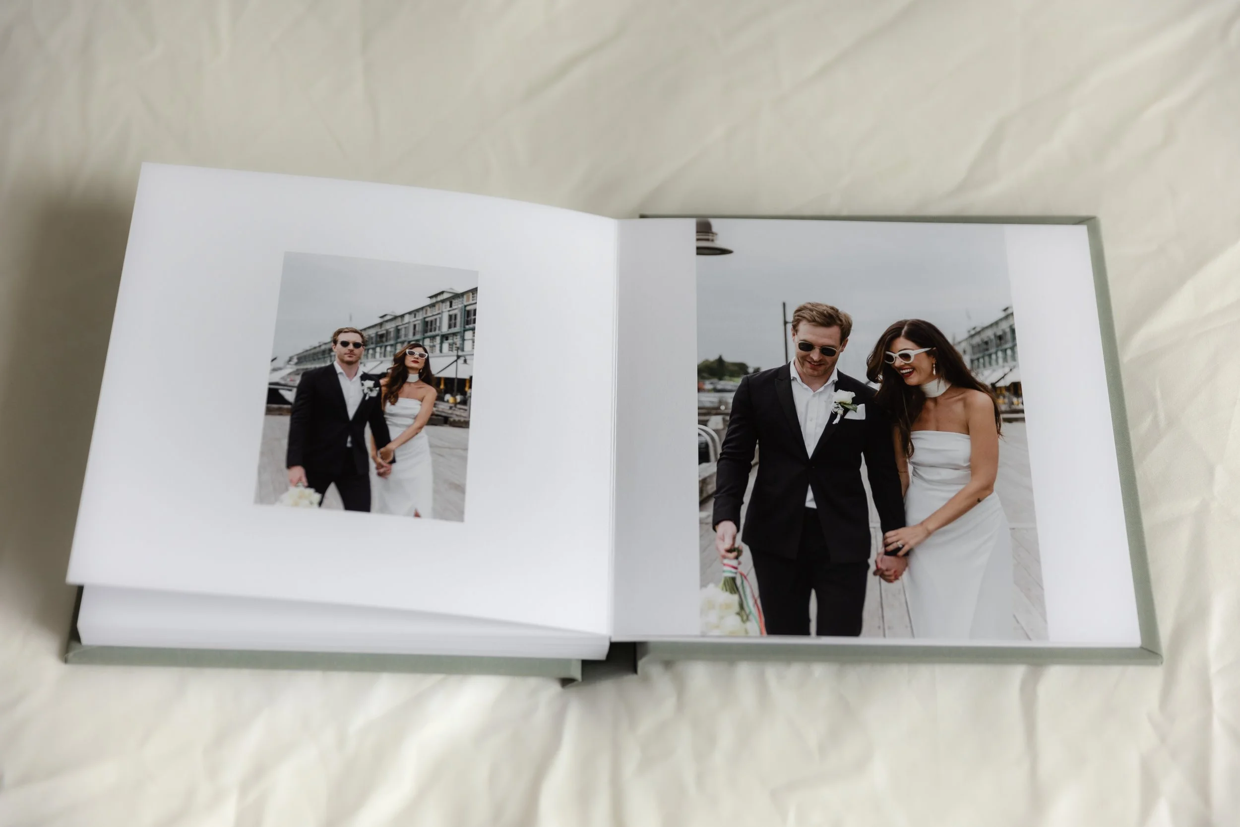 Open photo album with wedding photos, showing a newlywed couple dressed in wedding attire, holding hands and smiling outdoors near a harbor with boats and buildings in the background.