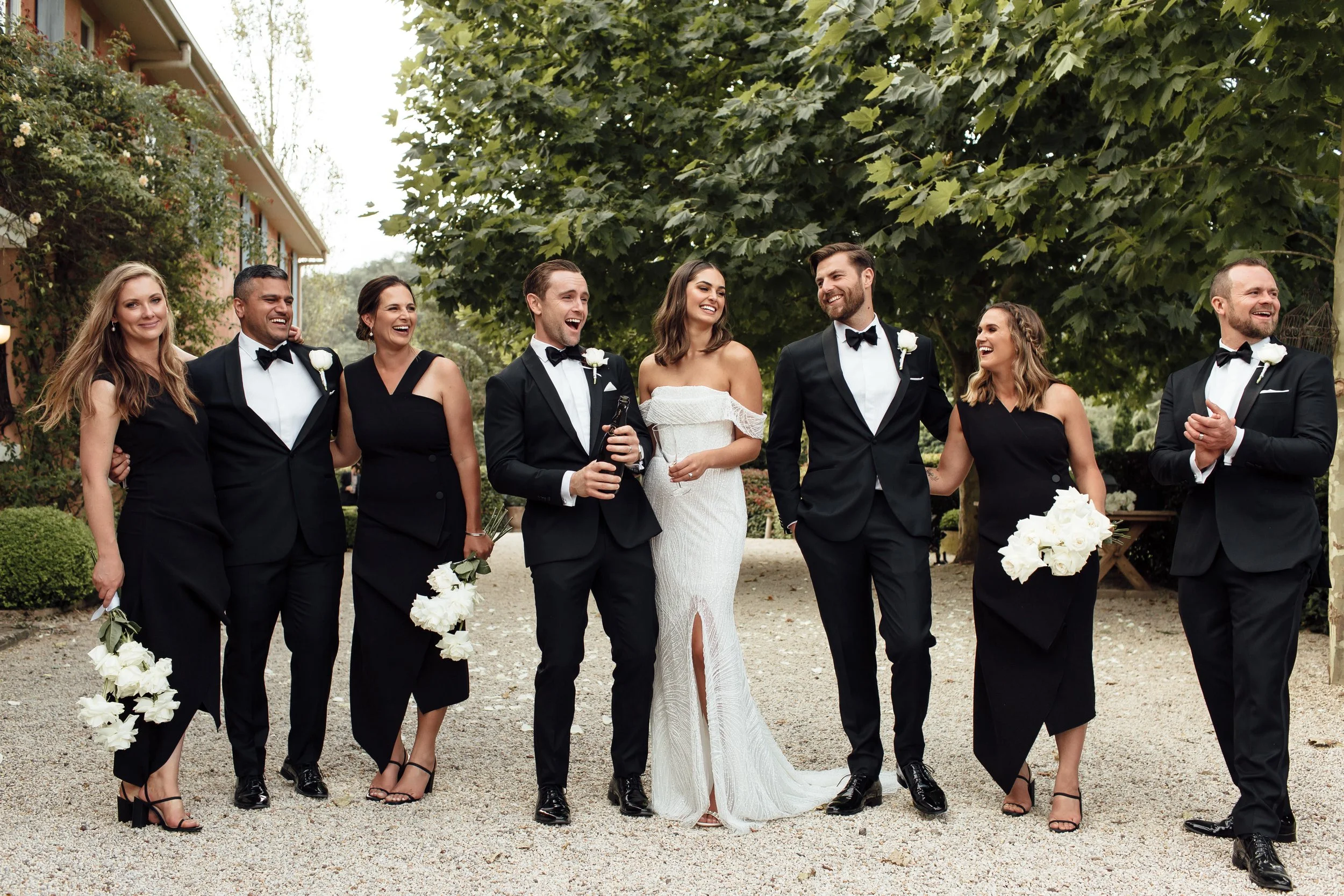 Wedding party, including bride and groom, standing outdoors at Redleaf Wollombi, smiling and laughing under a large leafy tree, with some holding white flower bouquets.