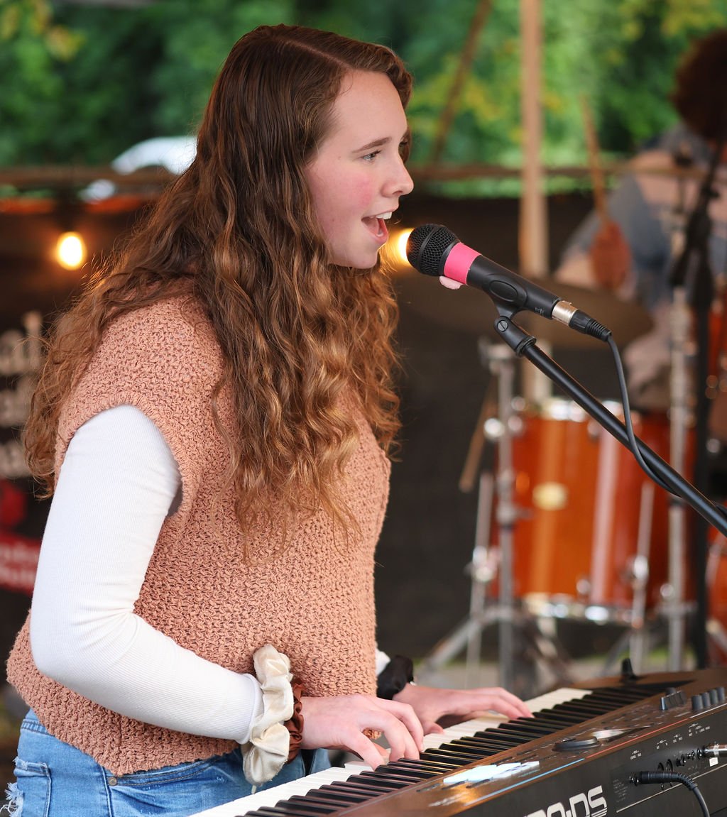 A girl with blonde hair and a sweater playing piano and singing in a rock band in Huntersville, NC